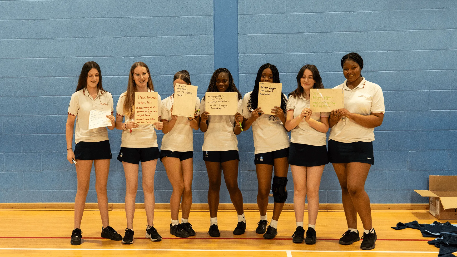 Seven girls in sports uniforms hold papers with handwritten clothing notes in a gym with blue walls.