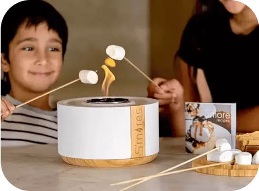 ילדים צולים מרשמלו על להבה במכשיר Smores במטבח מודרני ליד ספר מתכונים וקערה עם מרשמלו.