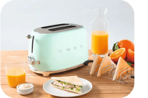 מטחן טוסט ירוק בהיר של SMEG על שולחן עץ עם מיץ תפוזים, כריך משולש, ותפוזים שלמים וחצויים.