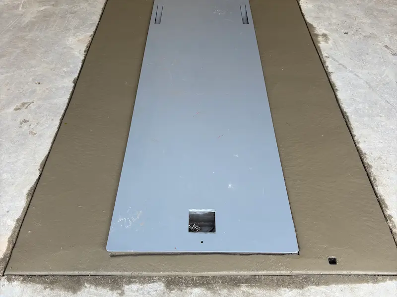 Gray metal plate covering a rectangular brown patch embedded in a light concrete floor.