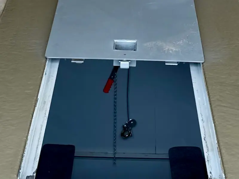 Partially open metal hatch door with a chain and hook hanging inside a dark space.