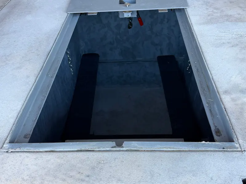 Open metal hatch in concrete floor revealing a small dark interior space with two long black cushions.