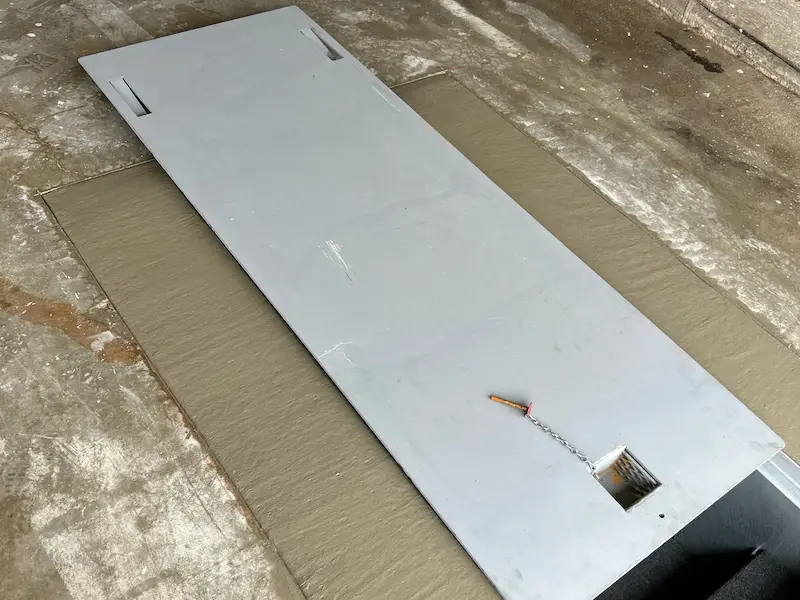 Large flat gray metal panel with cutouts lying on a textured concrete floor.