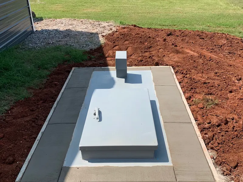 A rectangular gray metal hatch on a concrete slab surrounded by grass and red soil outside.