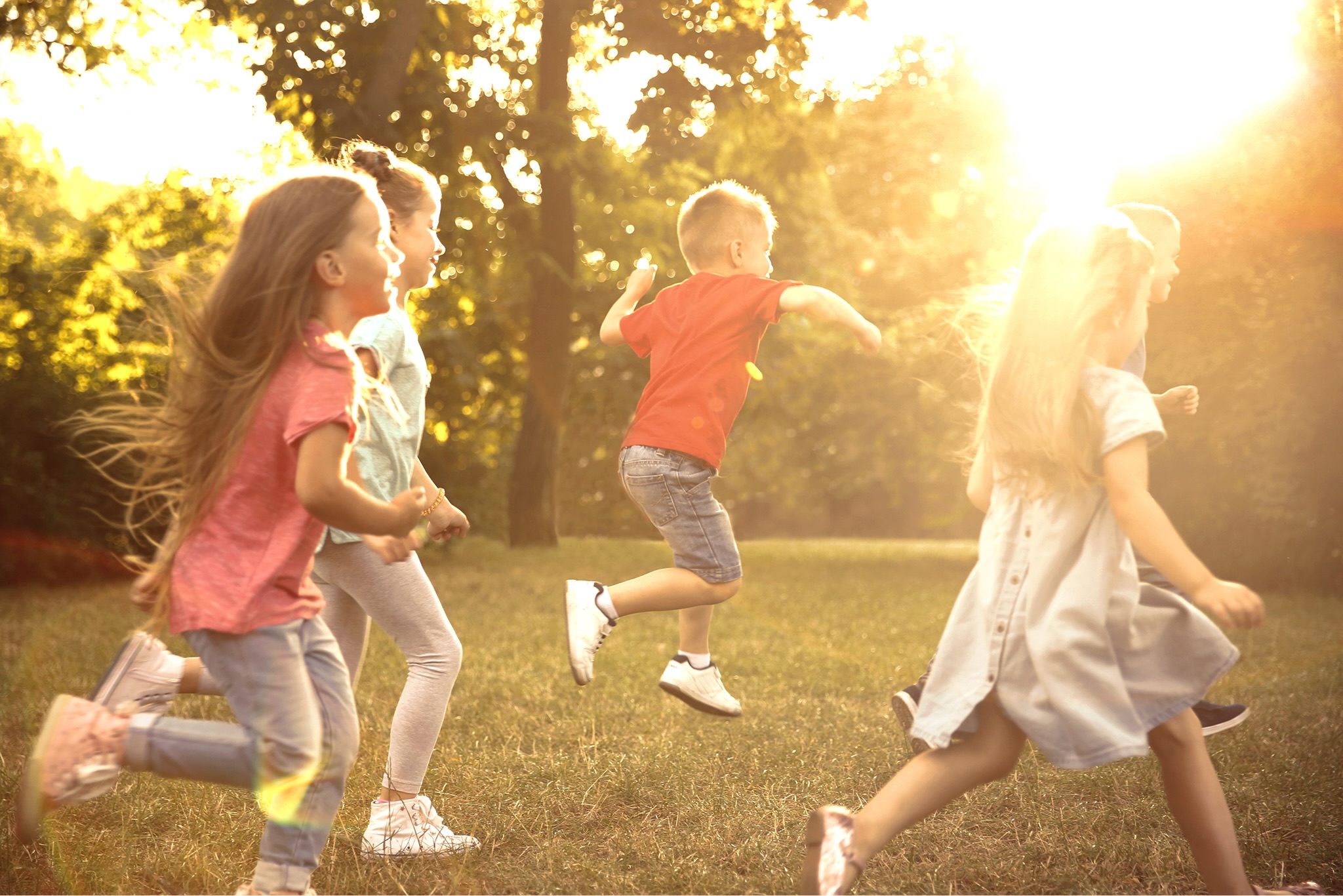 Children running and playing in a sunlit grassy field, golden-hour backlighting through trees in the background.