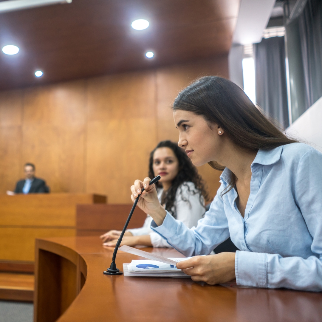 A woman with dark brown hair and a light blue long sleeved button down shirt testifying in court as another woman with curly dark hair and a white shirt looks on.