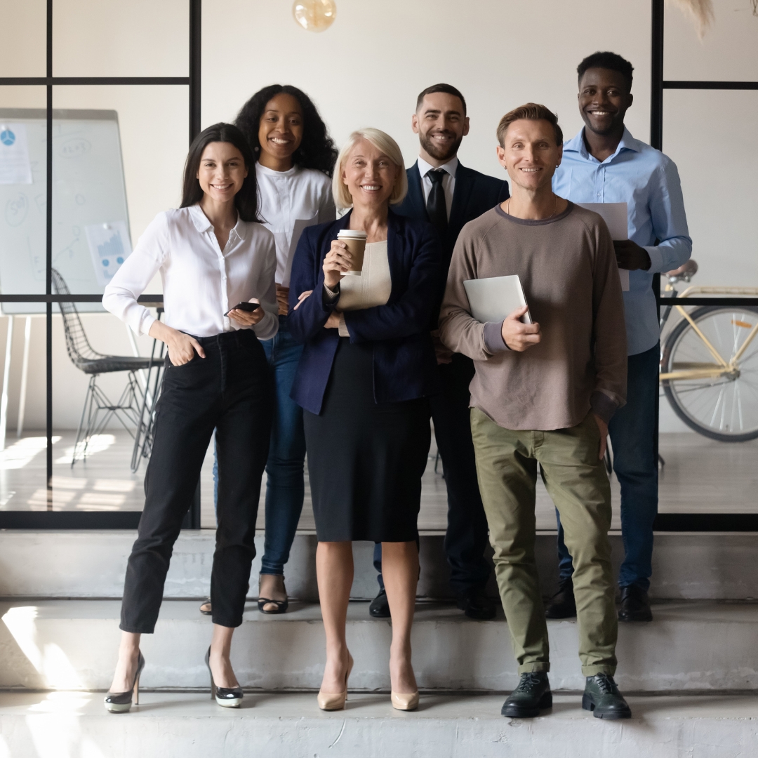 A multicultural group of six people smiling and standing in two rows. They are dressed in casual and professional attire and holding different objects in their hands.