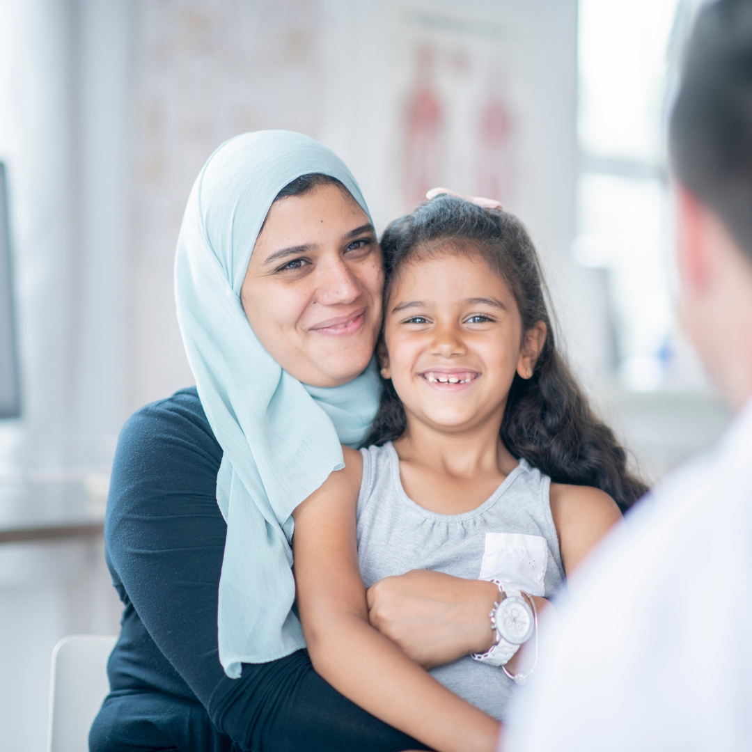 A woman with a light blue headscarf holding a girl in her arms and both are smiling and looking at the blurry silhouette of a healthcare provider