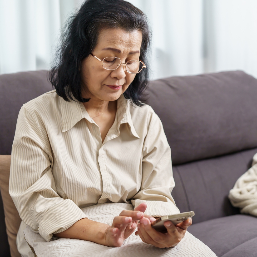 An older Asian woman in a beige dress and gold rimmed glasses looking at a mobile device