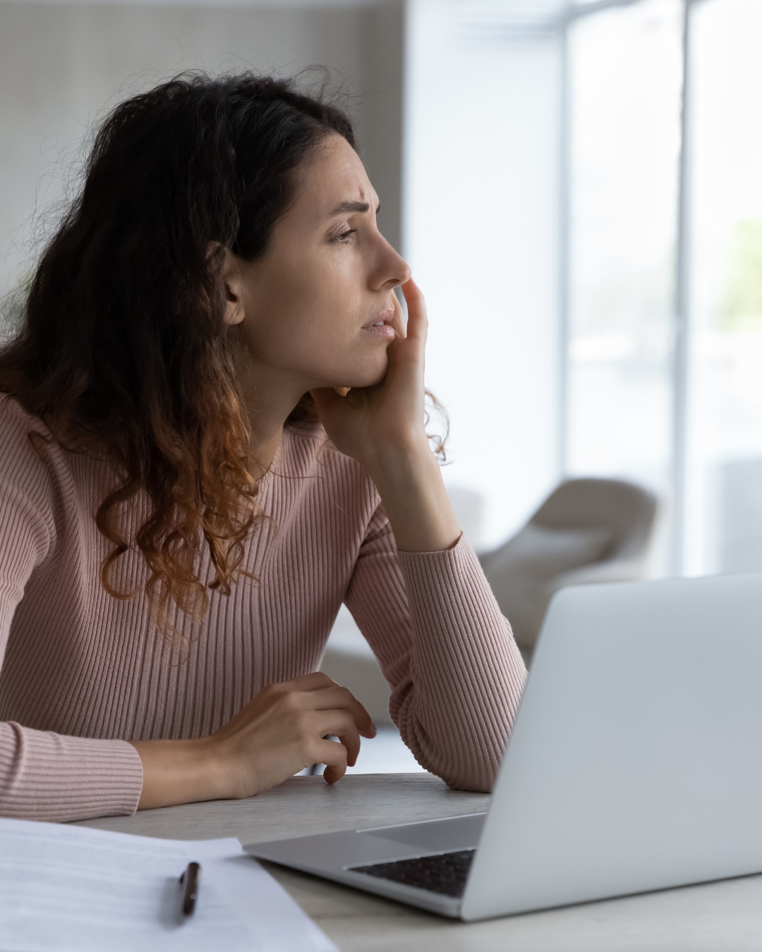 A woman with brown hair and a pink sweater sitting at a computer and looking off into the distance
