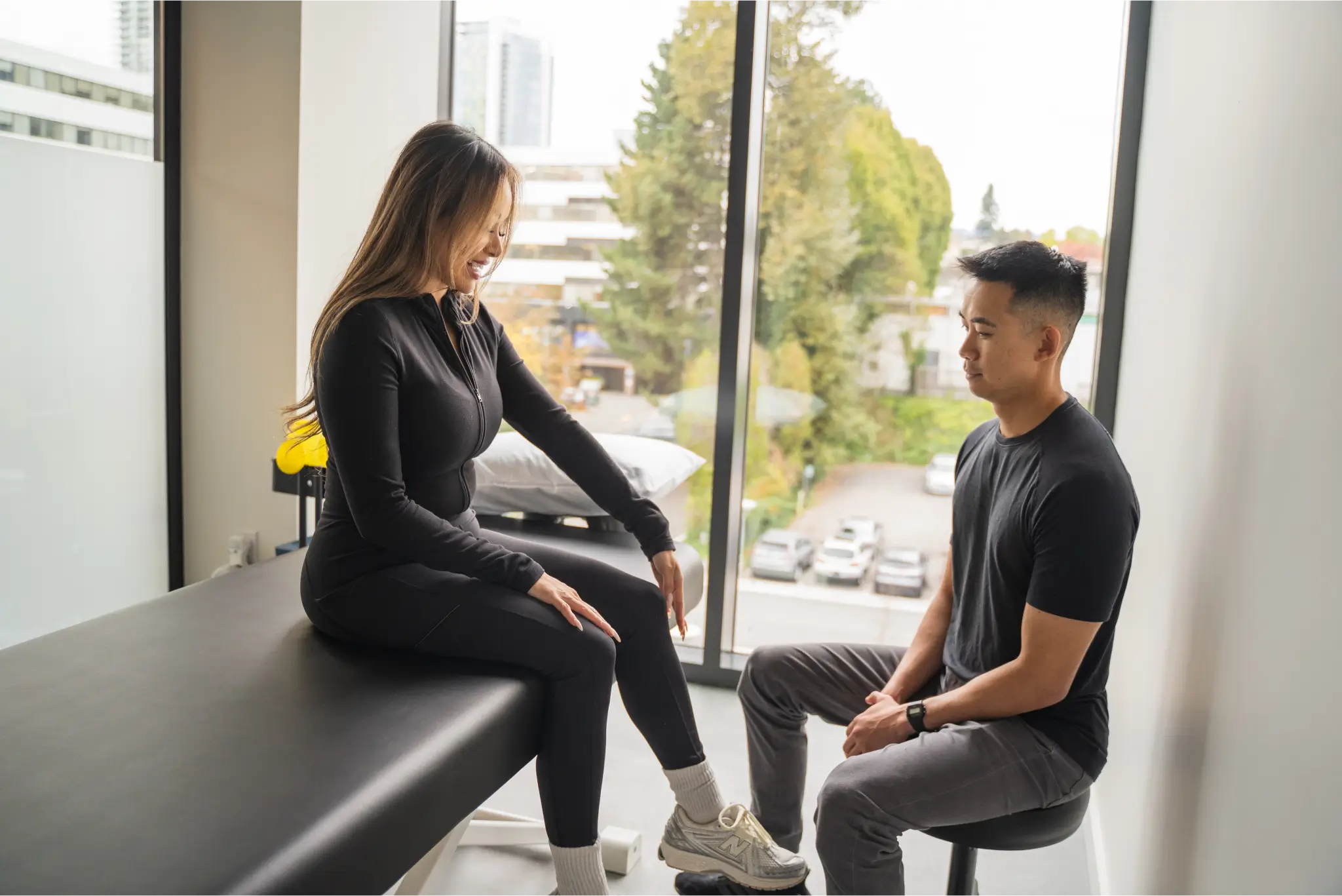 A physiotherapist talking a client through her knee assessment in a window-lit treatment room