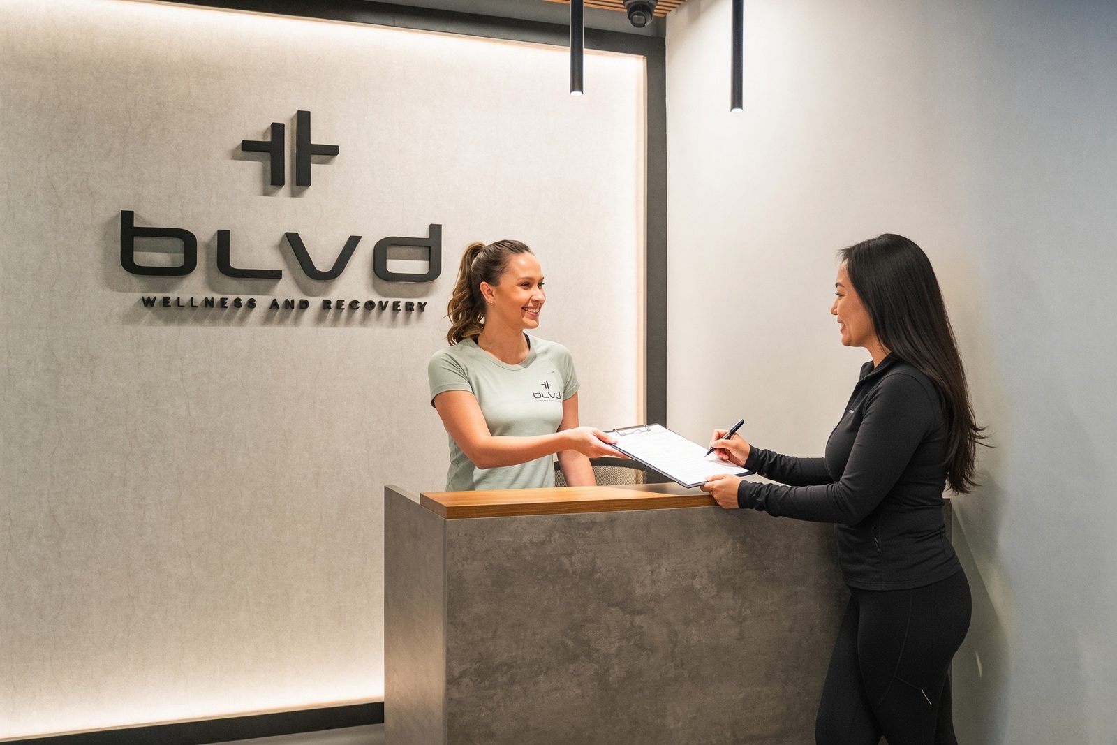 A BLVD team member handing a clipboard to a client at the reception desk, BLVD signage on the wall behind them