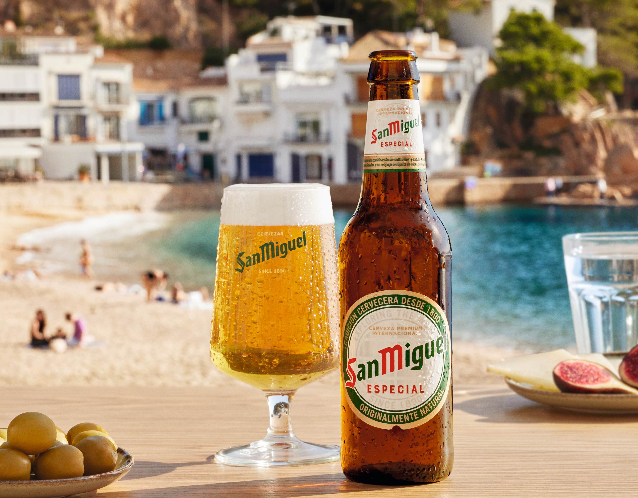 San Miquel bottle and glass competition campaign image on the table with olives and blurred image of the beach with people and building in the background