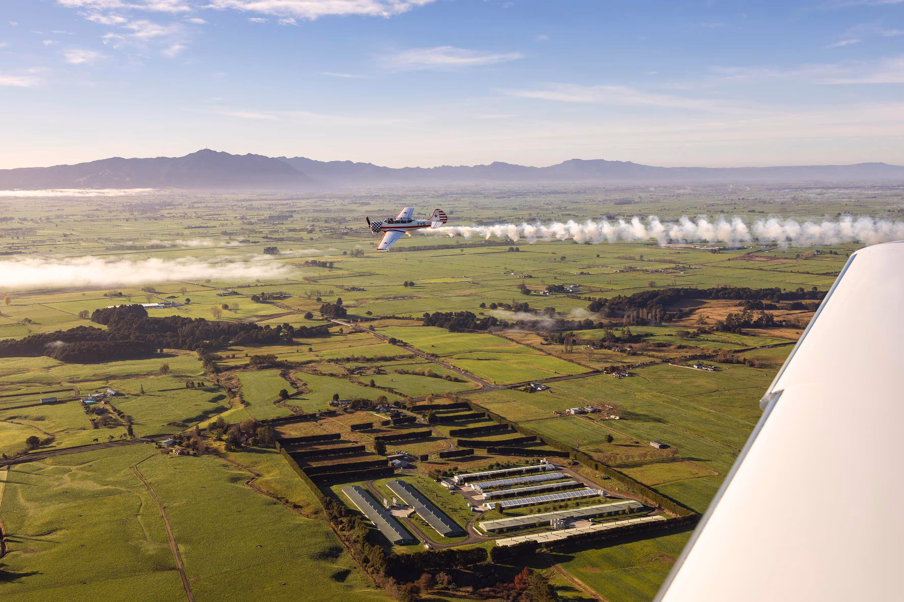 Aerial view of Te Kowhai Airpark showcasing a single aircraft performing acrobatics over the lush Waikato landscape. The image highlights the unique blend of aviation and residential living, with residential sections and hangars visible below.