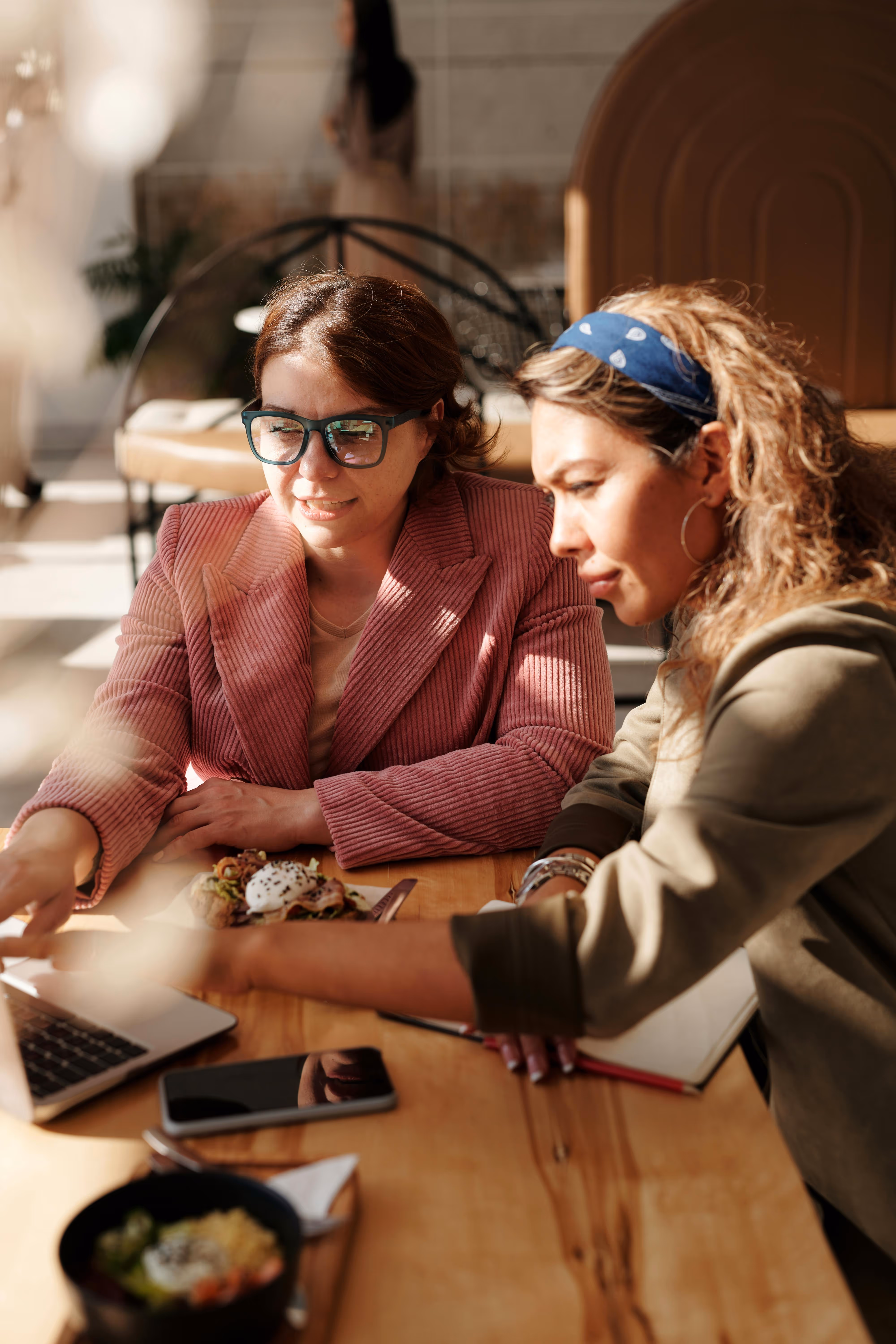 Two women engage in a collaborative discussion over a laptop in a cosy café setting. One woman points at the screen while the other observes attentively. Their table features notebooks and refreshments, reflecting a dynamic workspace atmosphere.