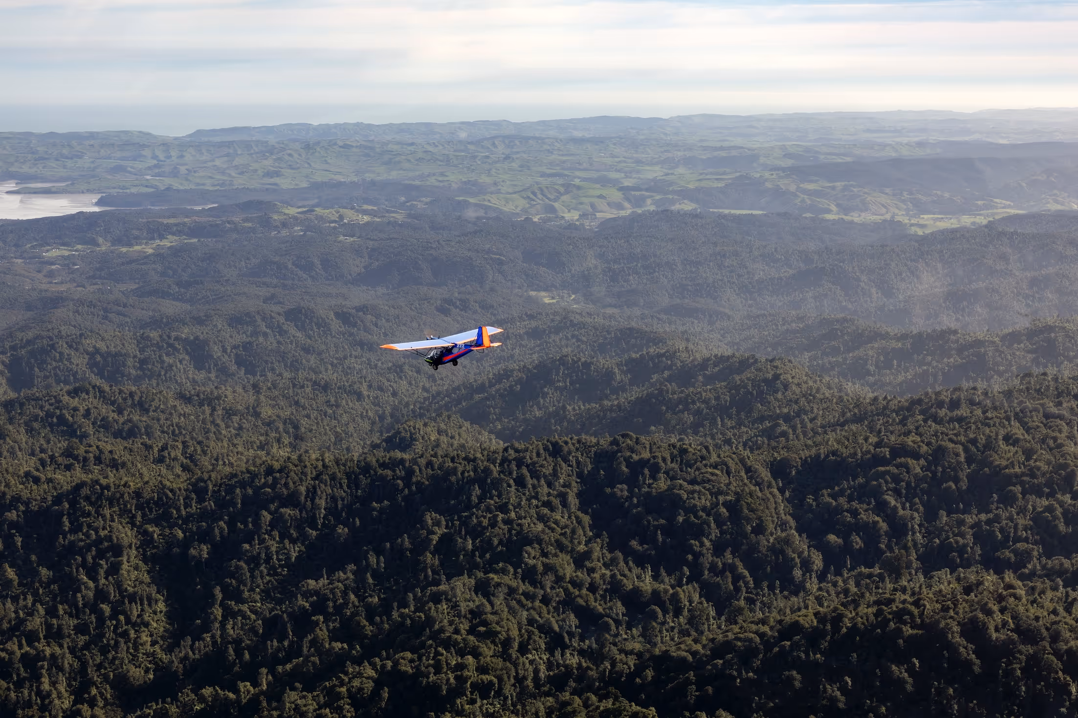 A small aircraft flying above the lush green hills of the Waikato region, showcasing the stunning natural landscape near Te Kowhai Airpark, New Zealand's first purpose-built aviation community, where aviation enthusiasts thrive.