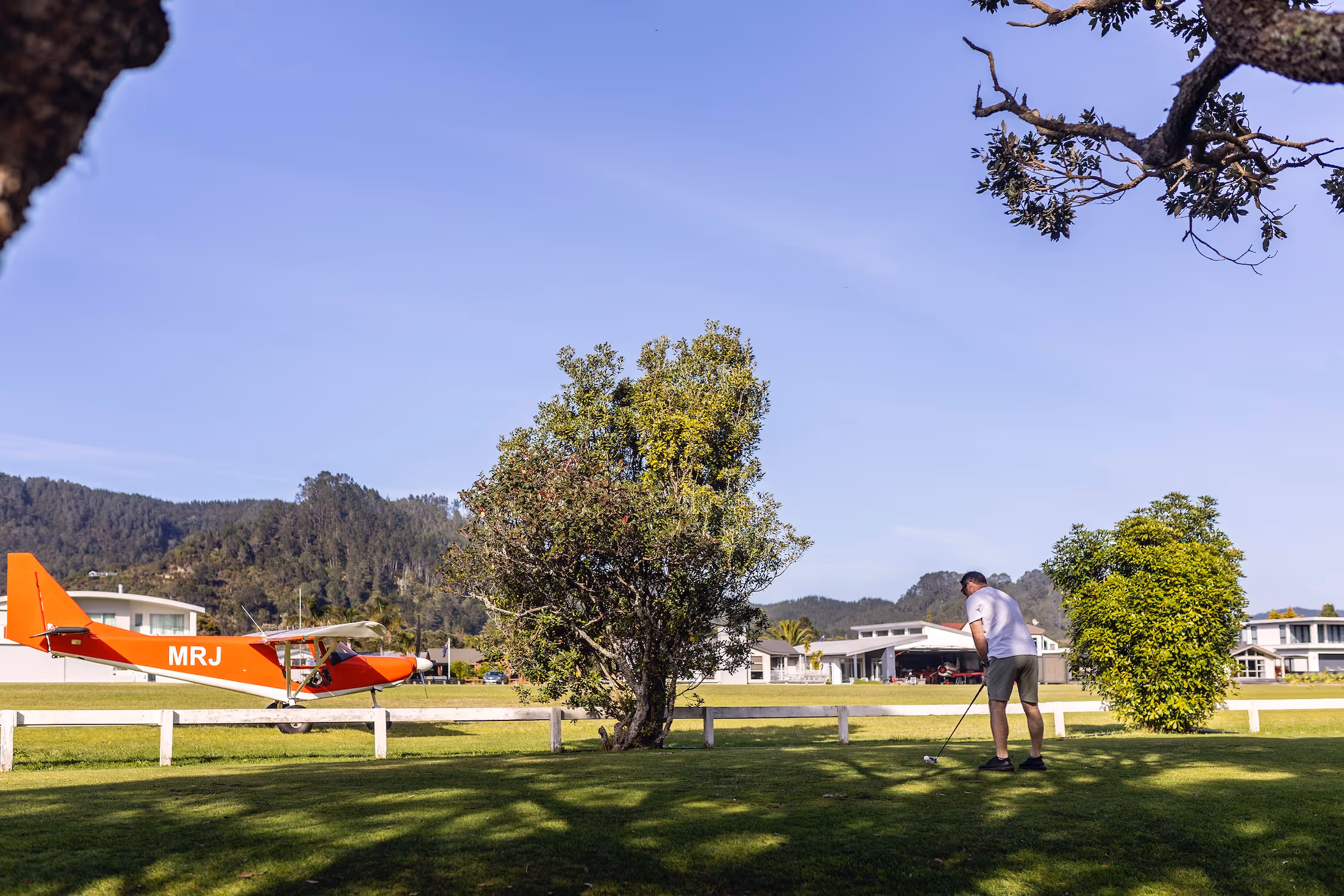 A vibrant scene at Te Kowhai Airpark, featuring a small orange aircraft on the grass, while a resident enjoys golfing nearby. The backdrop showcases modern homes and lush Waikato hills, highlighting this unique aviation community lifestyle.