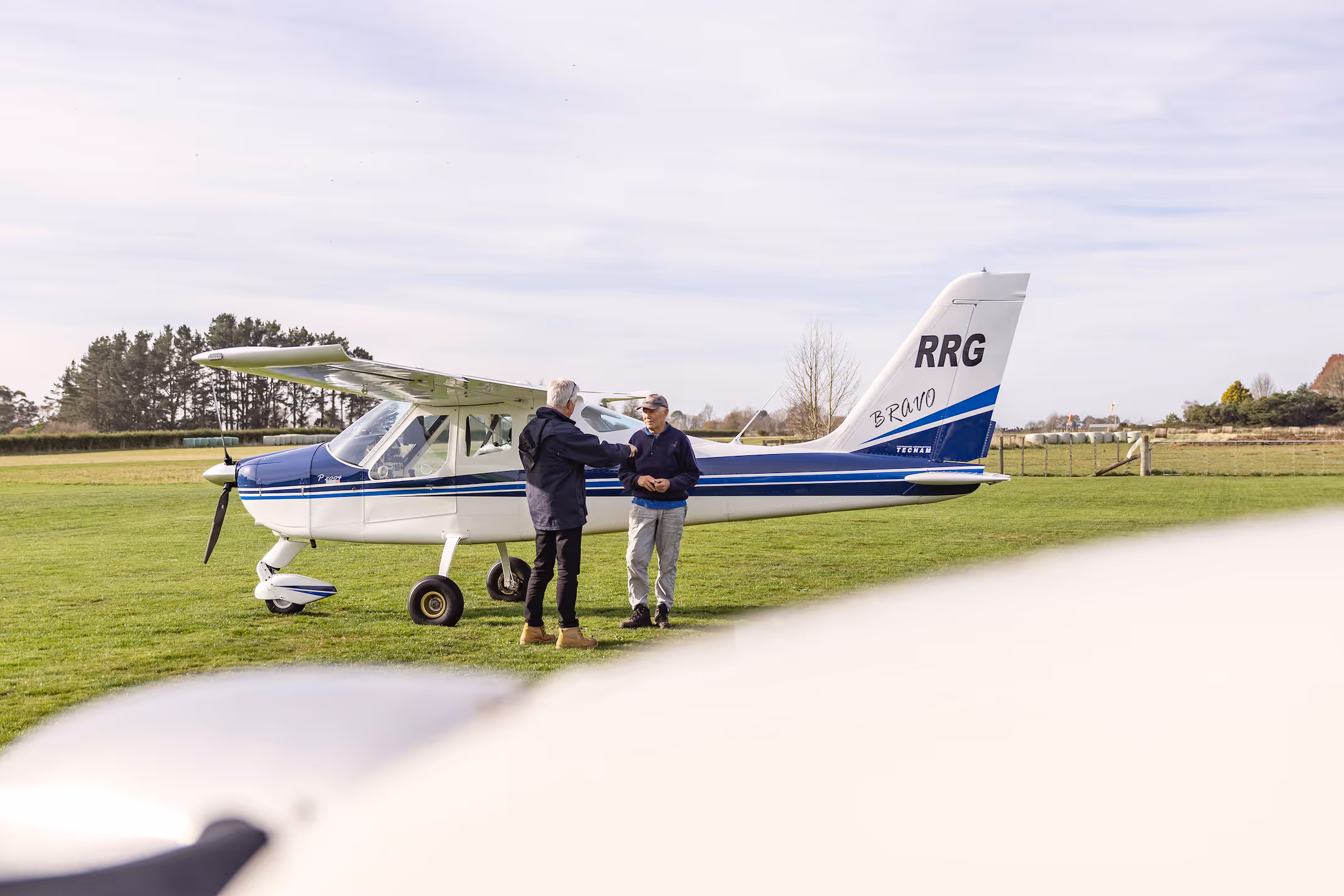 Two individuals shake hands next to a light aircraft on the lush grass at Te Kowhai Airpark, highlighting the unique aviation lifestyle. The picturesque Waikato landscape serves as a backdrop, emphasising community and direct airfield access.