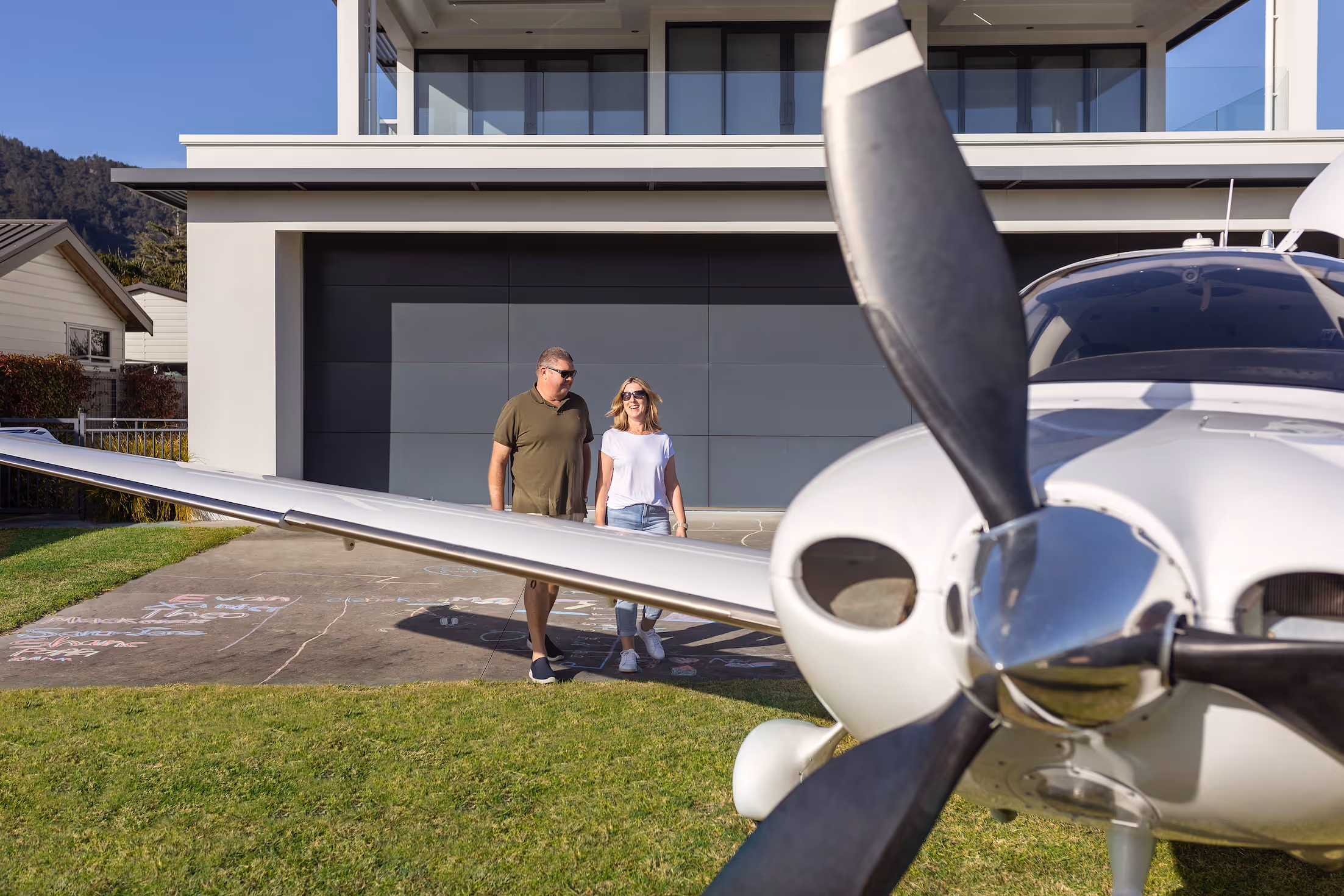 A couple strolls near their modern home with an attached hangar at Te Kowhai Airpark, showcasing the unique lifestyle of living with aircraft. The property features a private grass runway, blending residential comfort with aviation passion in the Waikato region.