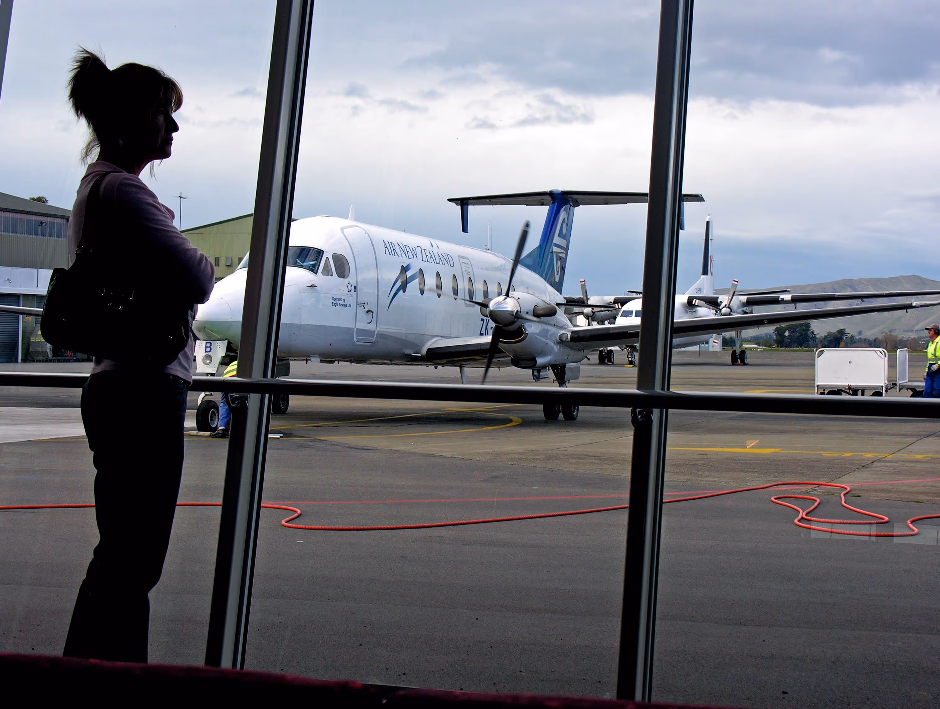 A silhouette of a woman stands at a terminal window, overlooking an Air New Zealand aircraft at the airport. This scene reflects the aviation lifestyle prominent in Te Kowhai Airpark, blending modern travel with an aviation community atmosphere.