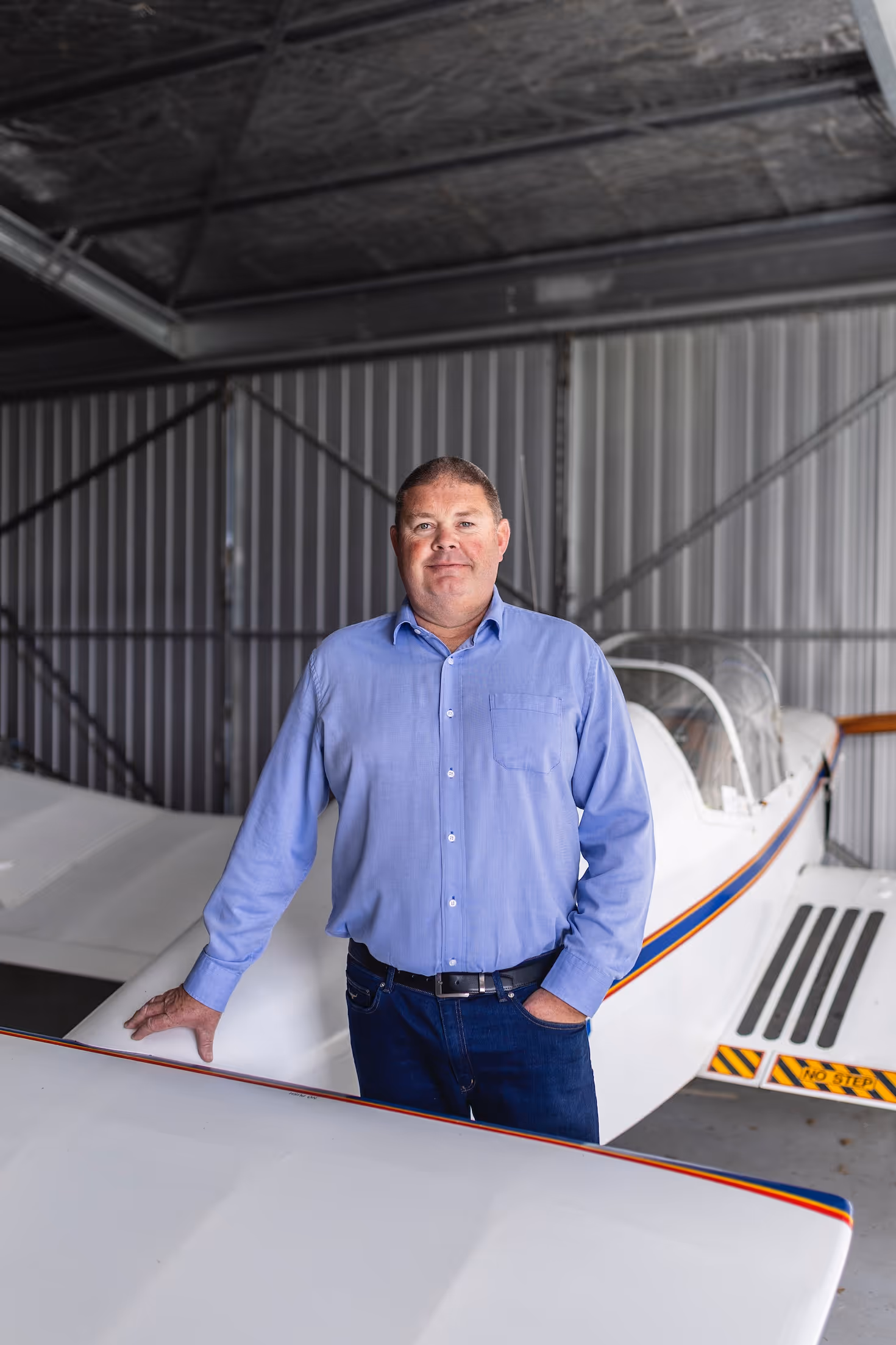 A smiling man stands confidently beside a small aircraft in a hangar at Te Kowhai Airpark, New Zealand’s first aviation community. The setting highlights the integration of residential living and aviation, showcasing a vibrant lifestyle for aviation enthusiasts.
