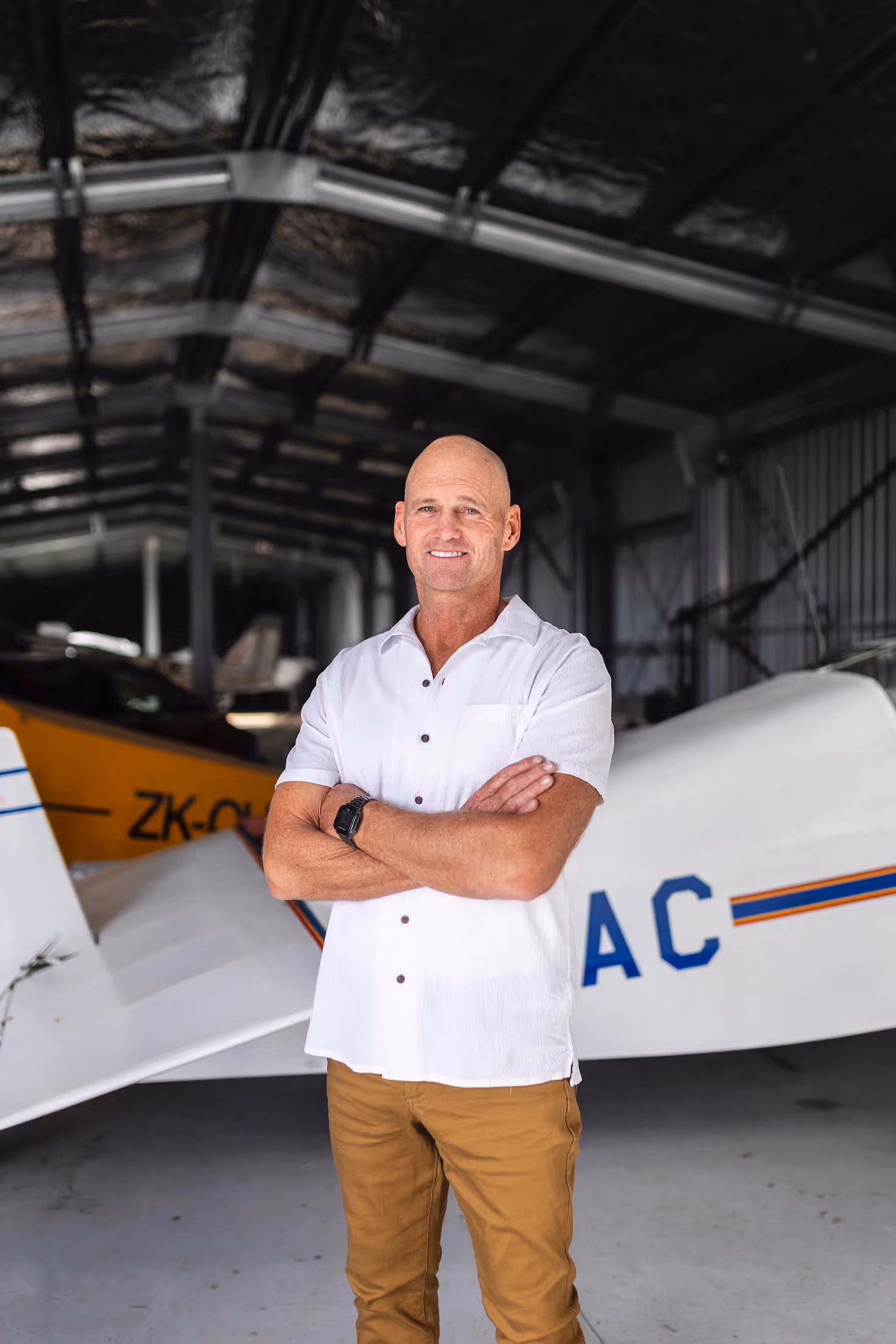Smiling man standing confidently with arms crossed in an aircraft hangar at Te Kowhai Airpark, New Zealand. The background features a vintage aircraft, highlighting the unique aviation community lifestyle integrated with residential living nearby Hamilton.