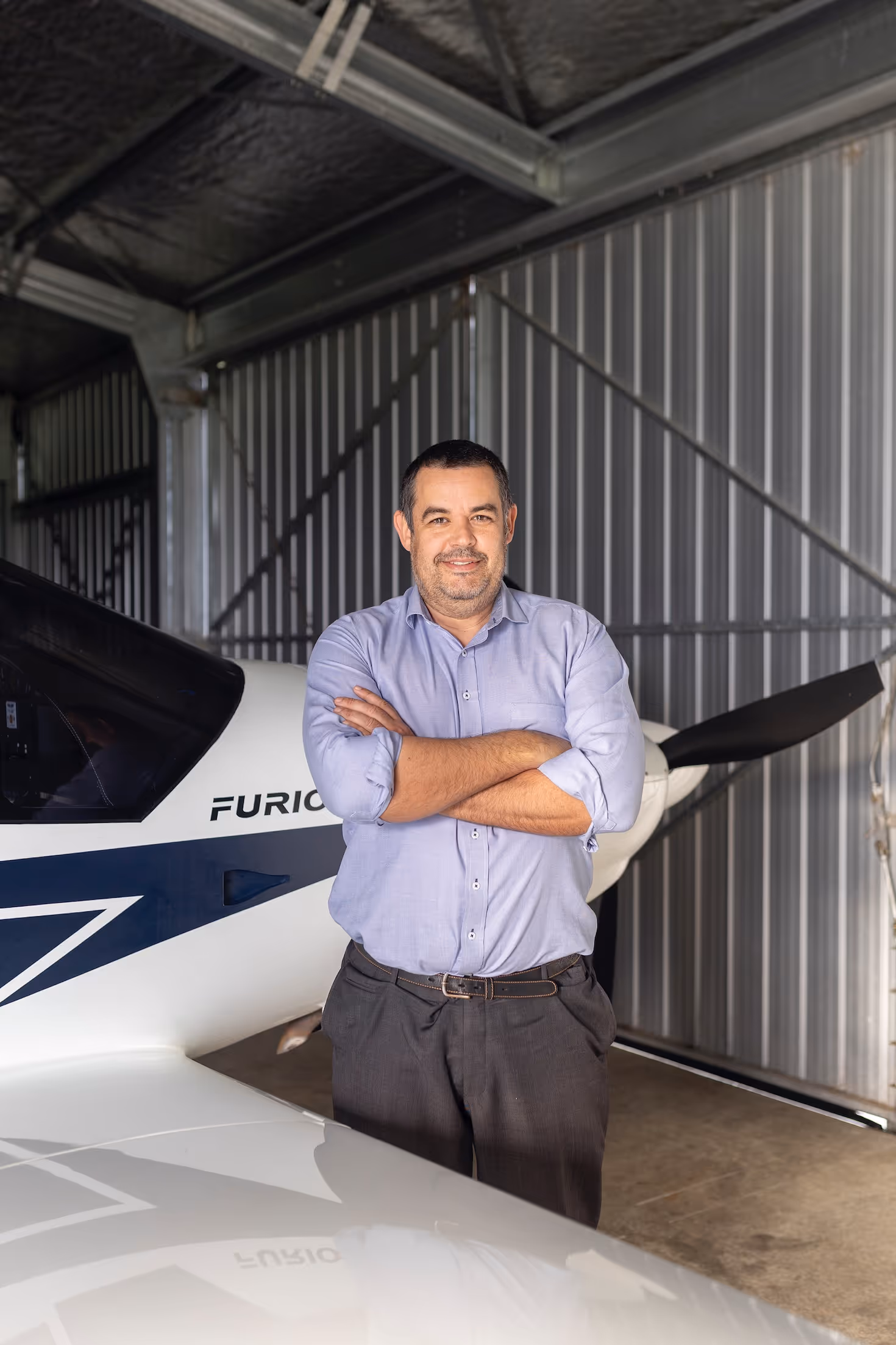 A confident man stands with crossed arms in a hangar at Te Kowhai Airpark, New Zealand. Behind him, a modern aircraft is visible, highlighting the unique lifestyle of aviation enthusiasts in this vibrant residential aviation community.