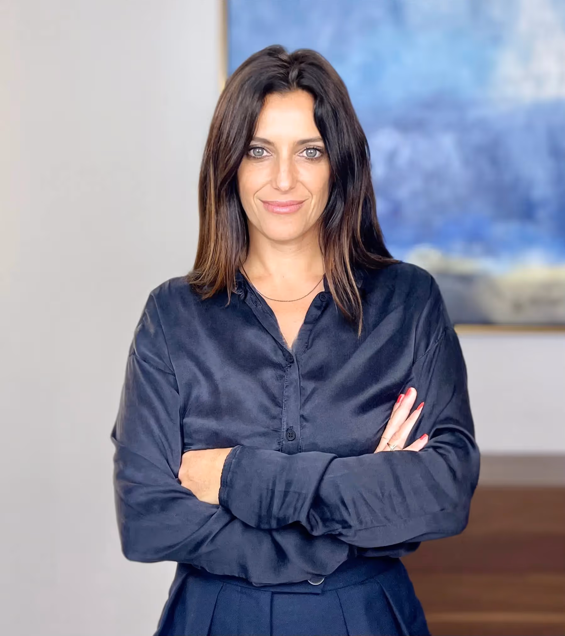 Woman with brown hair with black shirt, arms crossed, in front of a light wall with a blurred blue board.