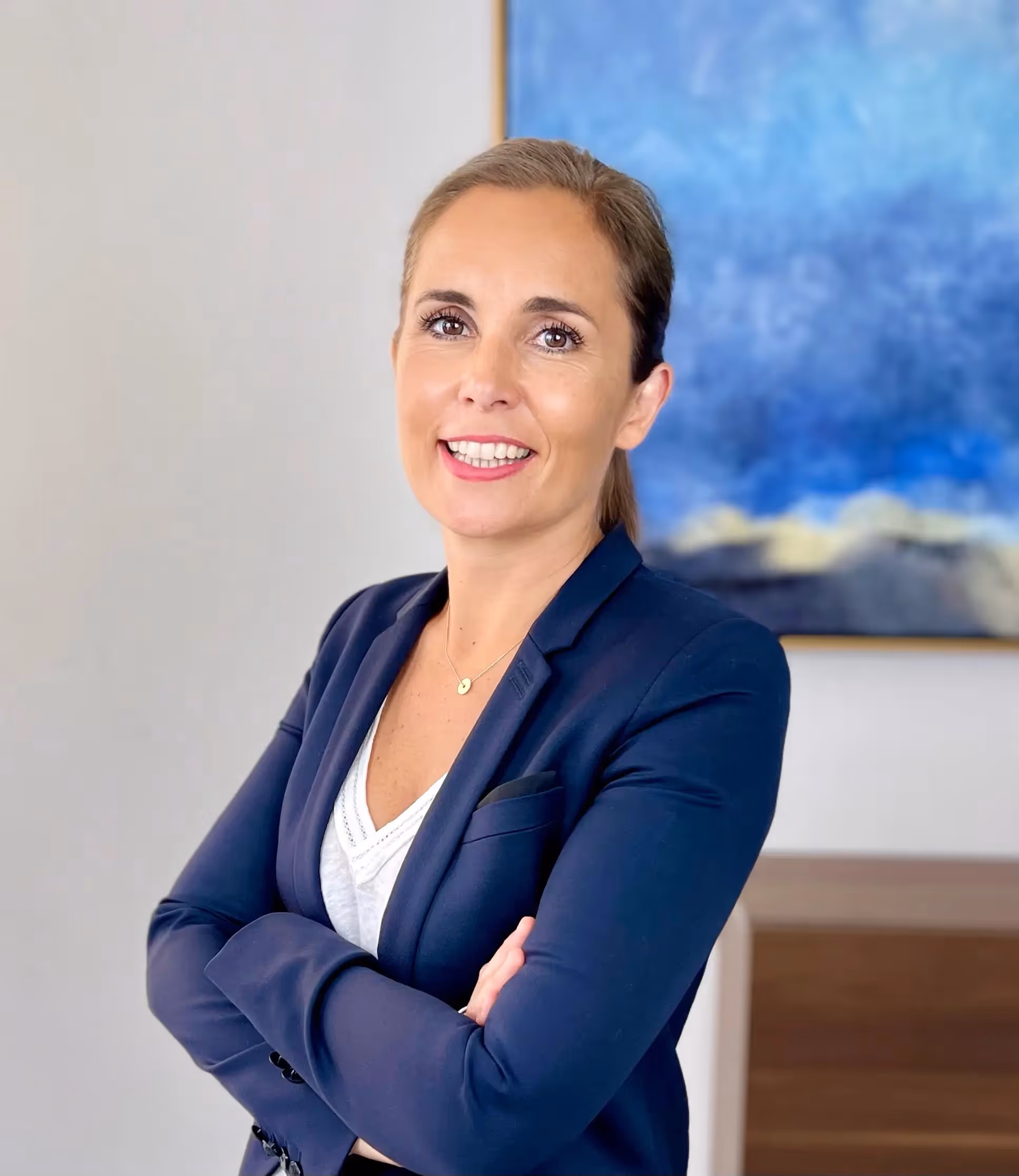 Smiling woman in a navy blue jacket with her arms crossed in front of a light wall and a blue abstract painting in the background.
