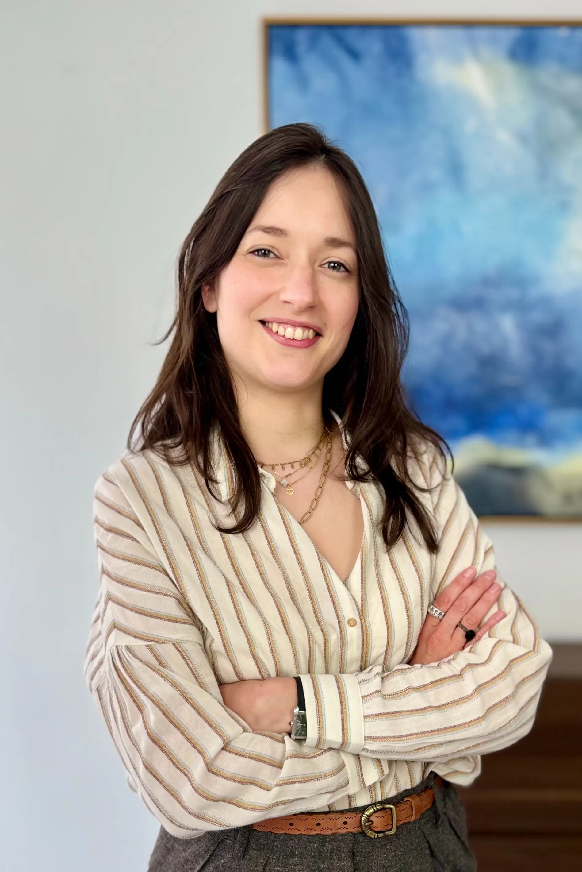 A smiling woman with brown hair, wearing a beige striped shirt with her arms crossed, in front of a blurry blue board.