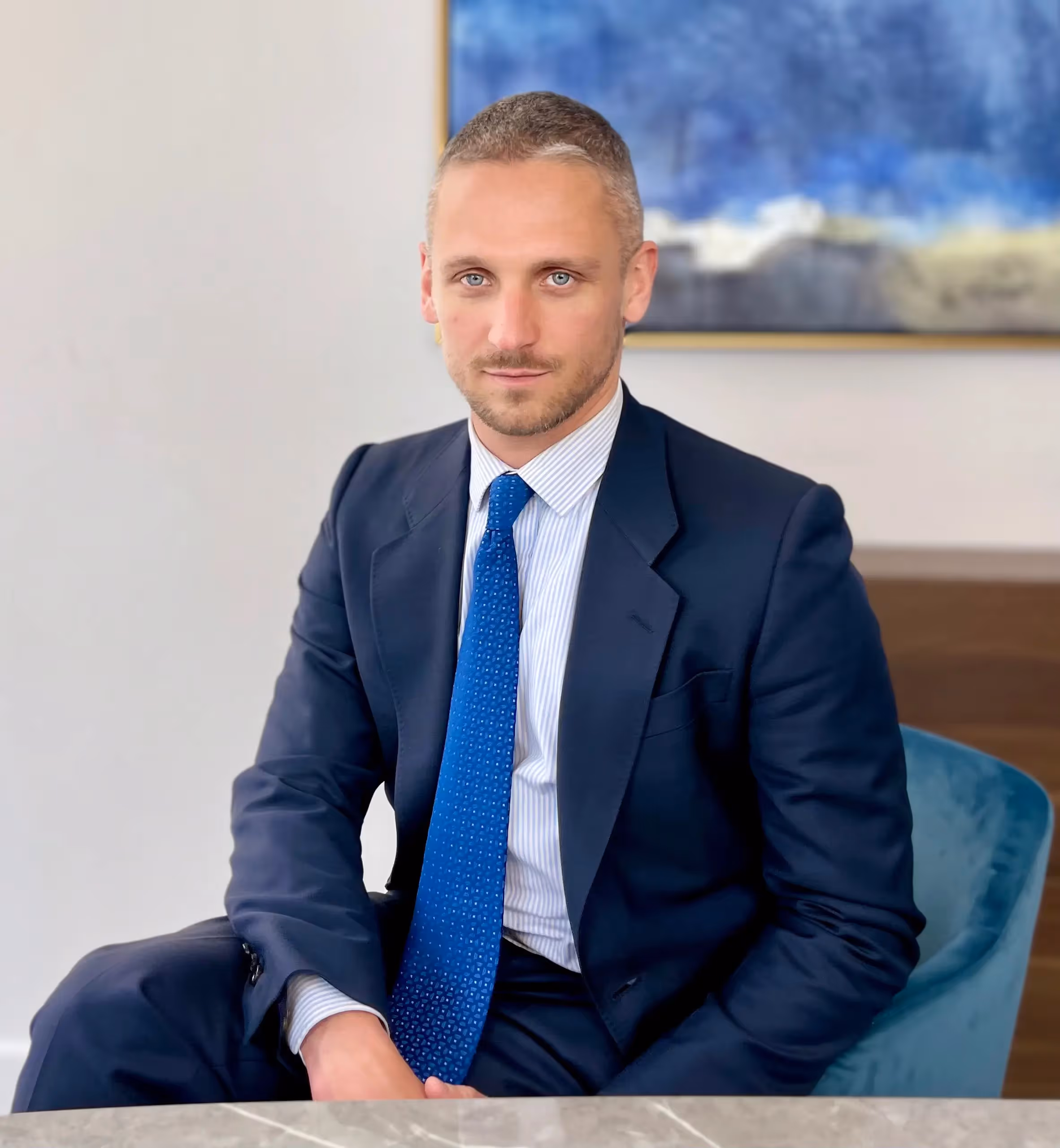 Man in a navy suit with blue tie sitting on a blue chair in front of a marble table, with an abstract painting in the background.
