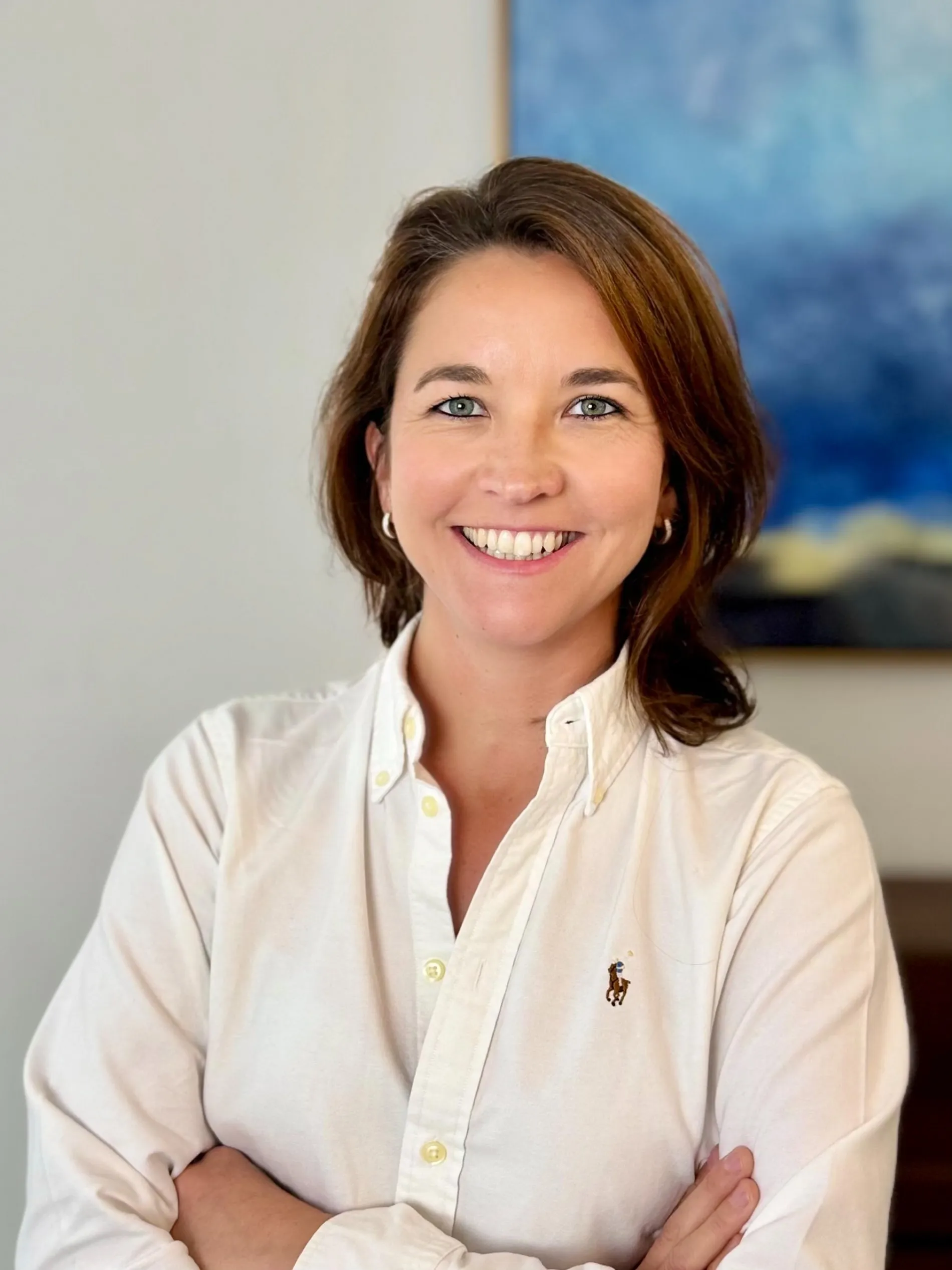 Smiling woman with brown hair, wearing a white long sleeve shirt, with her arms crossed.