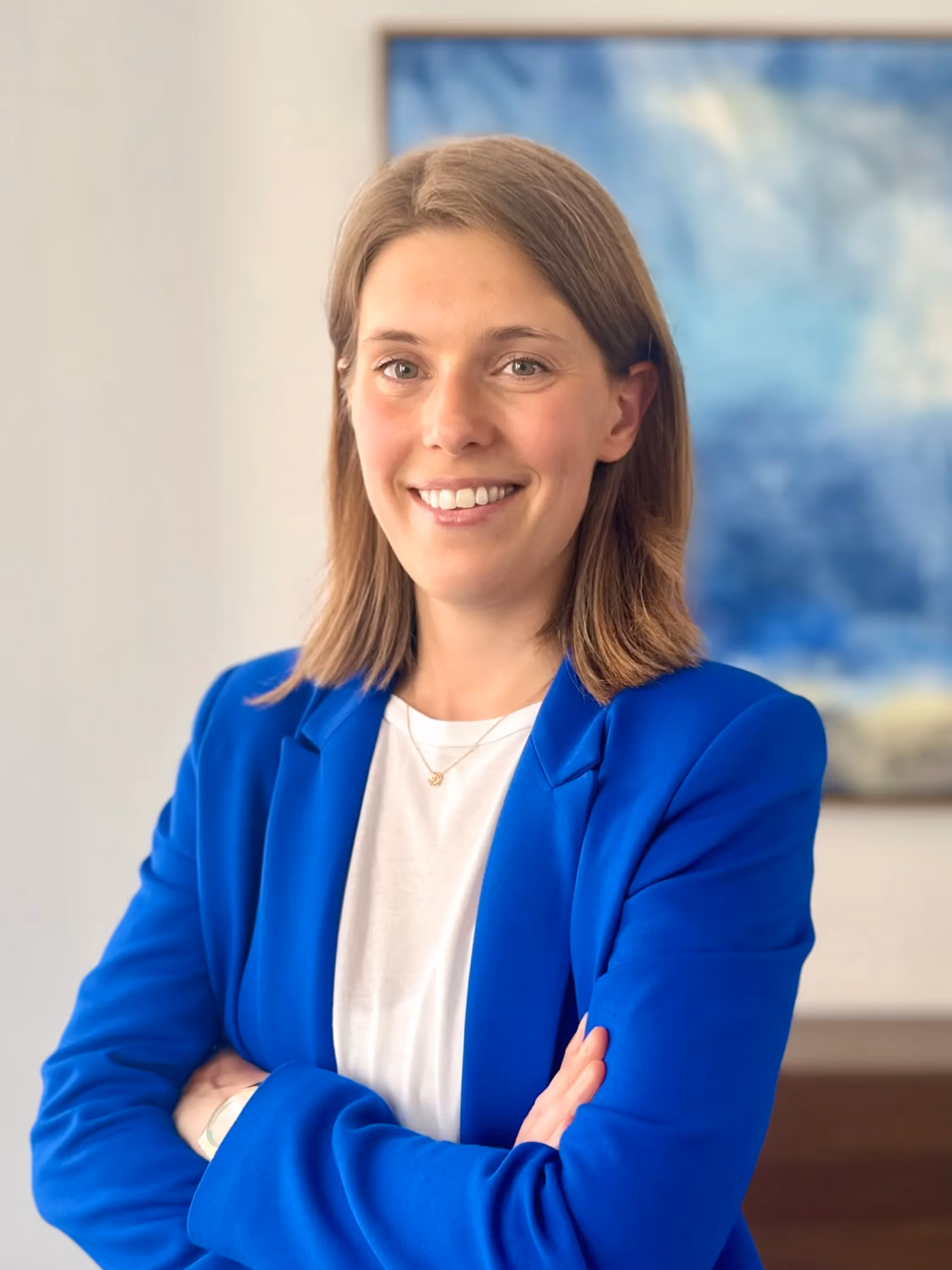 Smiling young woman with medium hair wearing a blue blazer and a white T-shirt, arms crossed, in front of a bright wall with a blurred blue board in the background.