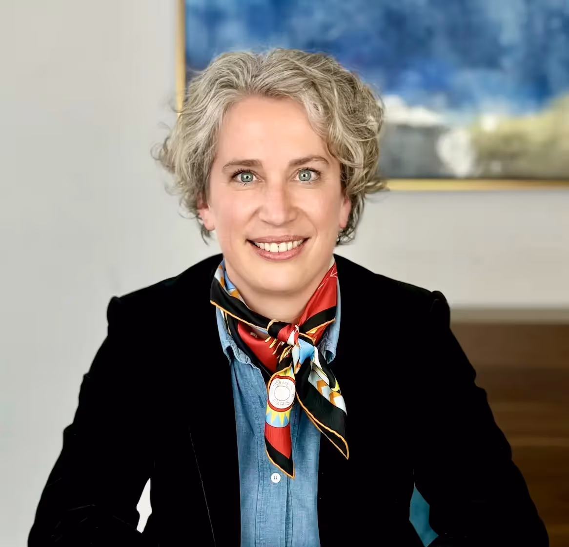 Portrait of a smiling woman with gray curly short hair, wearing a denim shirt, a black blazer, and a colorful scarf tied around her neck.