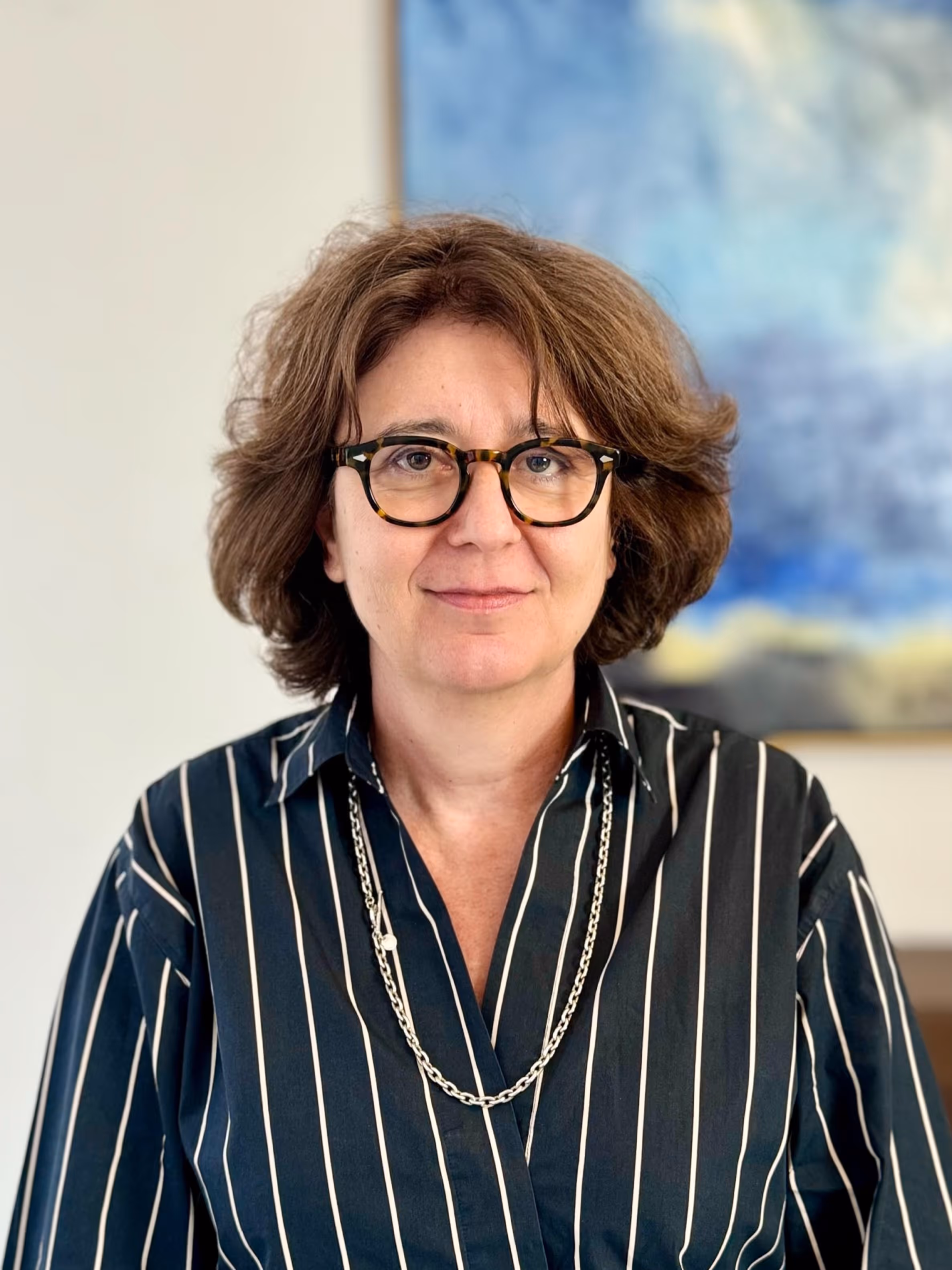 Woman with short curly hair wearing tortoiseshell glasses and a black shirt with white stripes and a silver necklace.