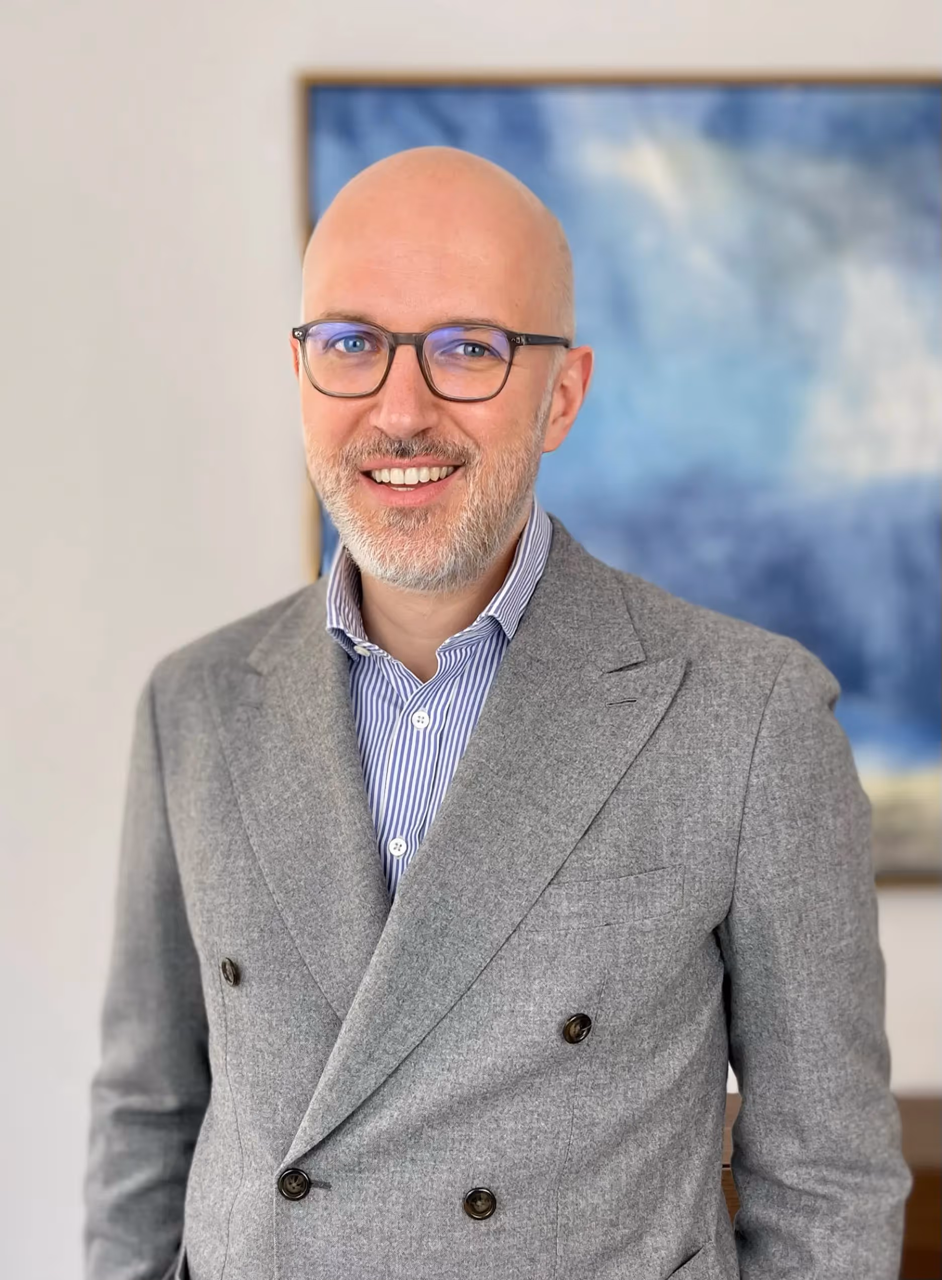 Bald man wearing glasses and a gray suit with striped shirt, smiling in front of a blurry blue paint.