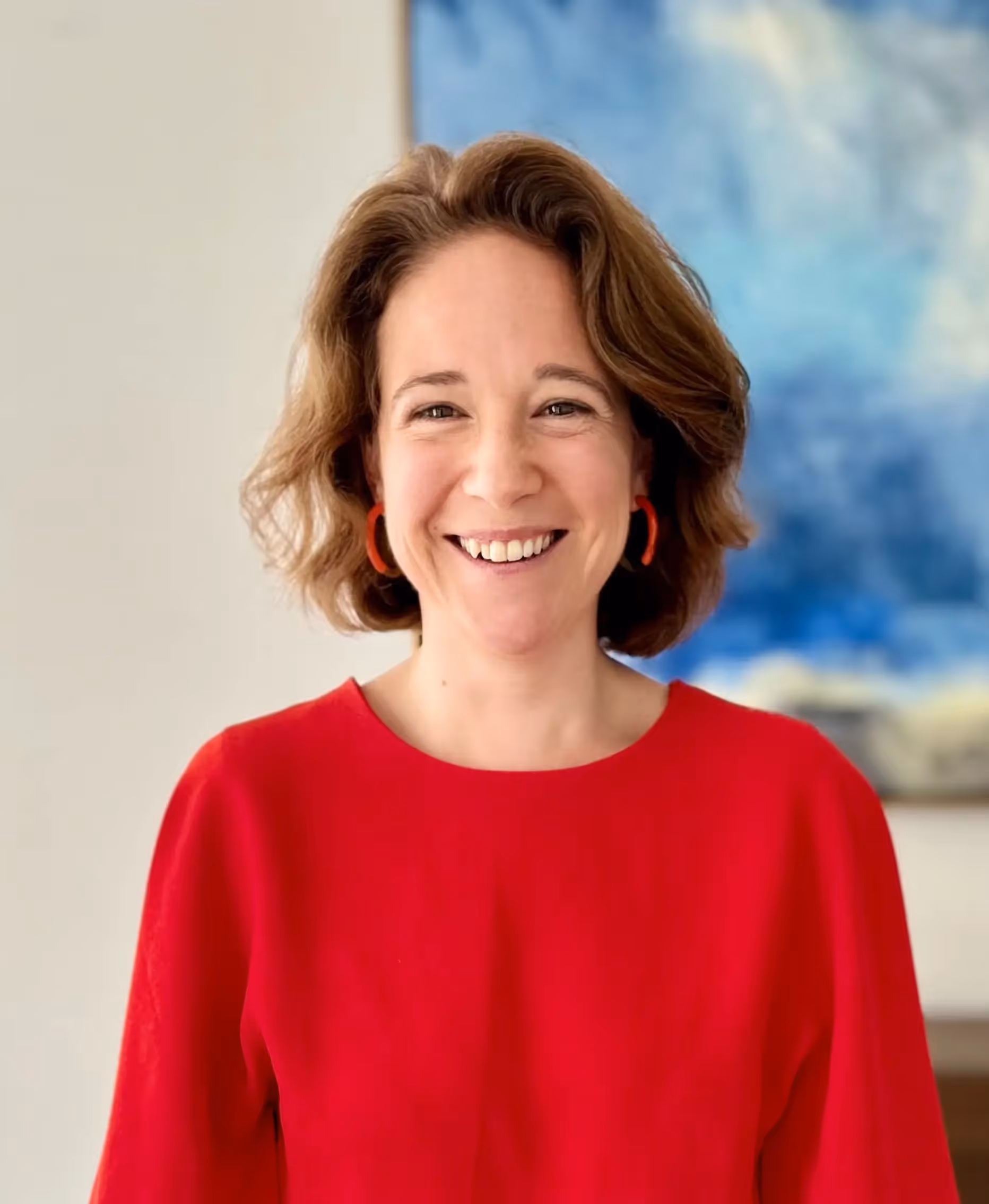 Smiling woman wearing a red sweater and red earrings, in front of a blue and white blurred background.
