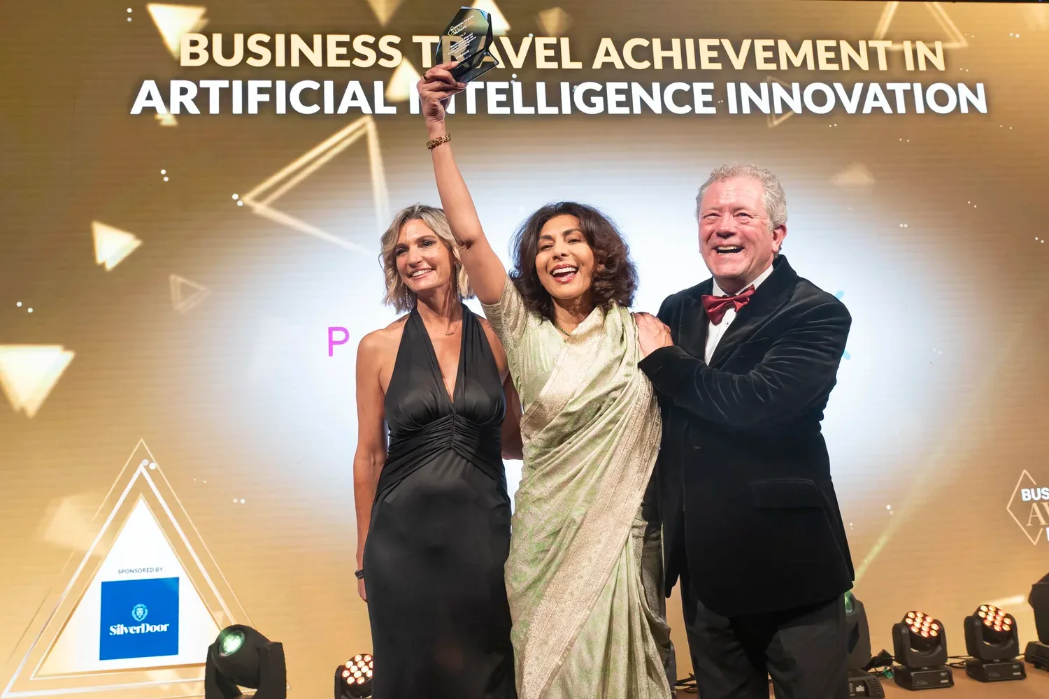A person in a pale green sari laughs, trophy raised high, between two people in black, a SilverDoor sponsor panel at left