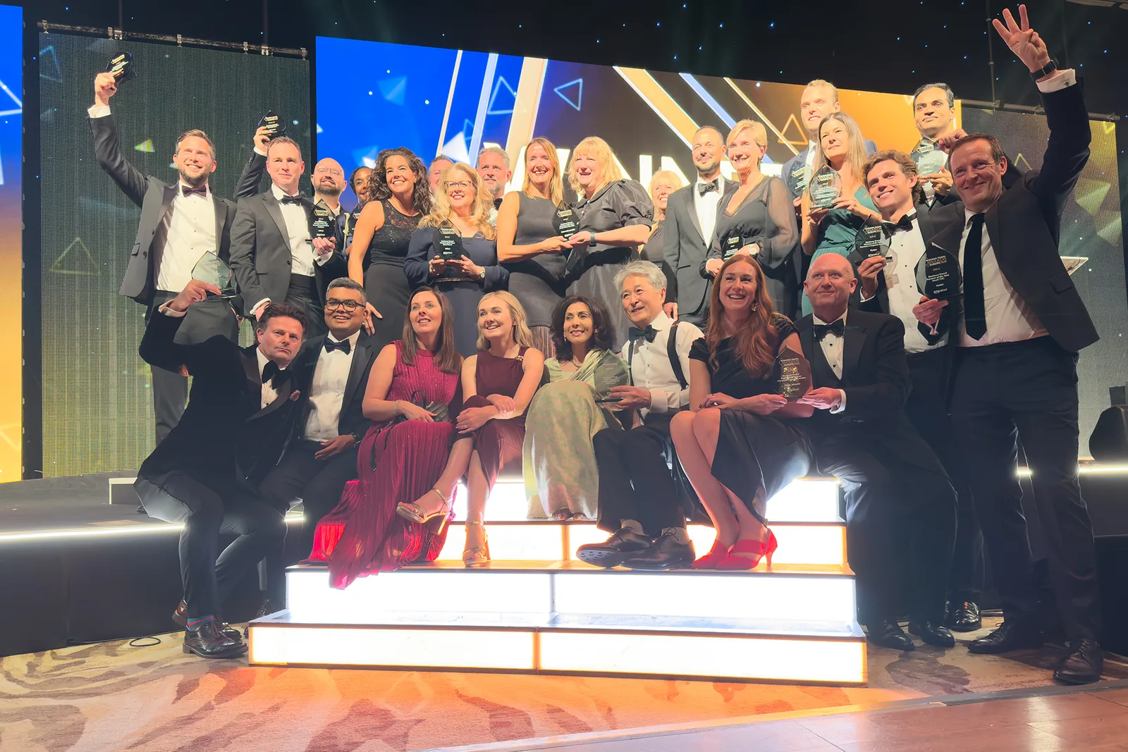 More than two dozen people in evening dress crowd lit stage steps, many holding glass trophies, a blue and gold screen behind