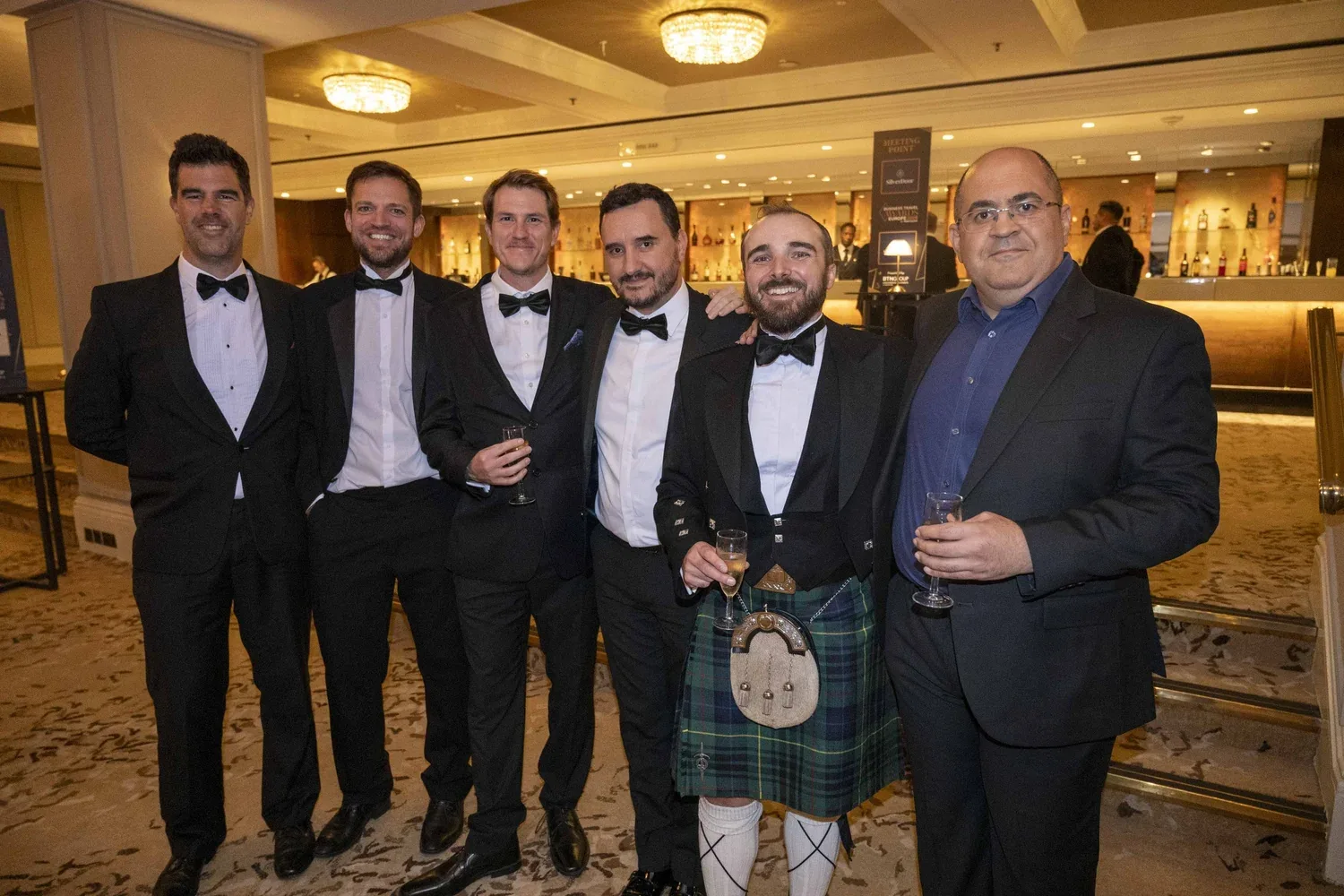 A row of men, most in black tie, stand close in a hotel bar, glasses in hand, chandeliers and lit bottles behind them