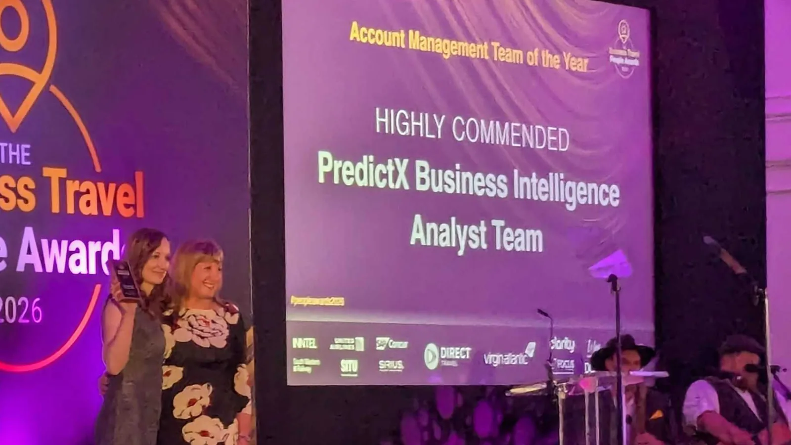 The screen reads highly commended, PredictX Business Intelligence Analyst Team; two people stand beside it, one with a plaque
