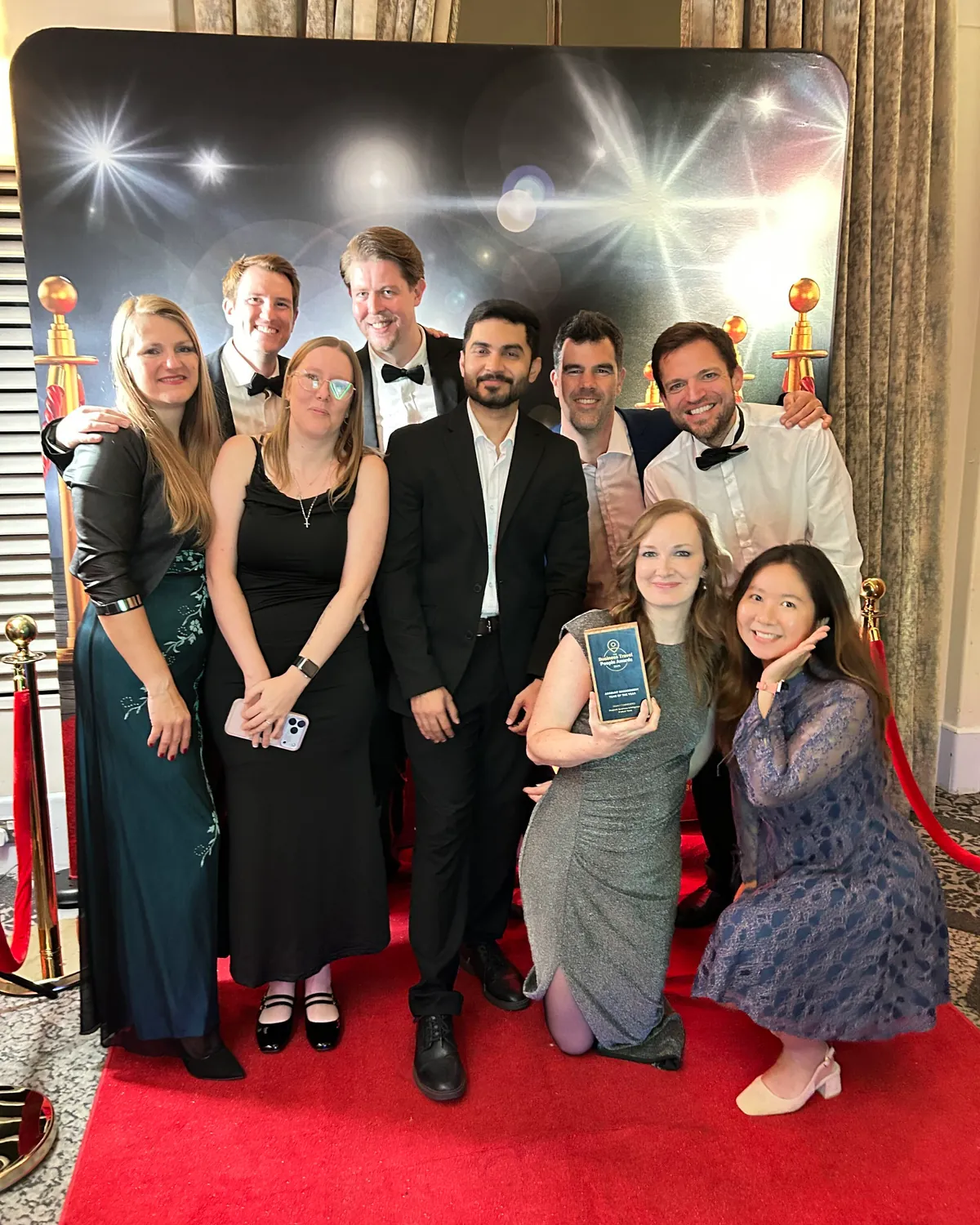 The same nine colleagues pose between gold rope posts before a starburst backdrop, the highly commended plaque at the front