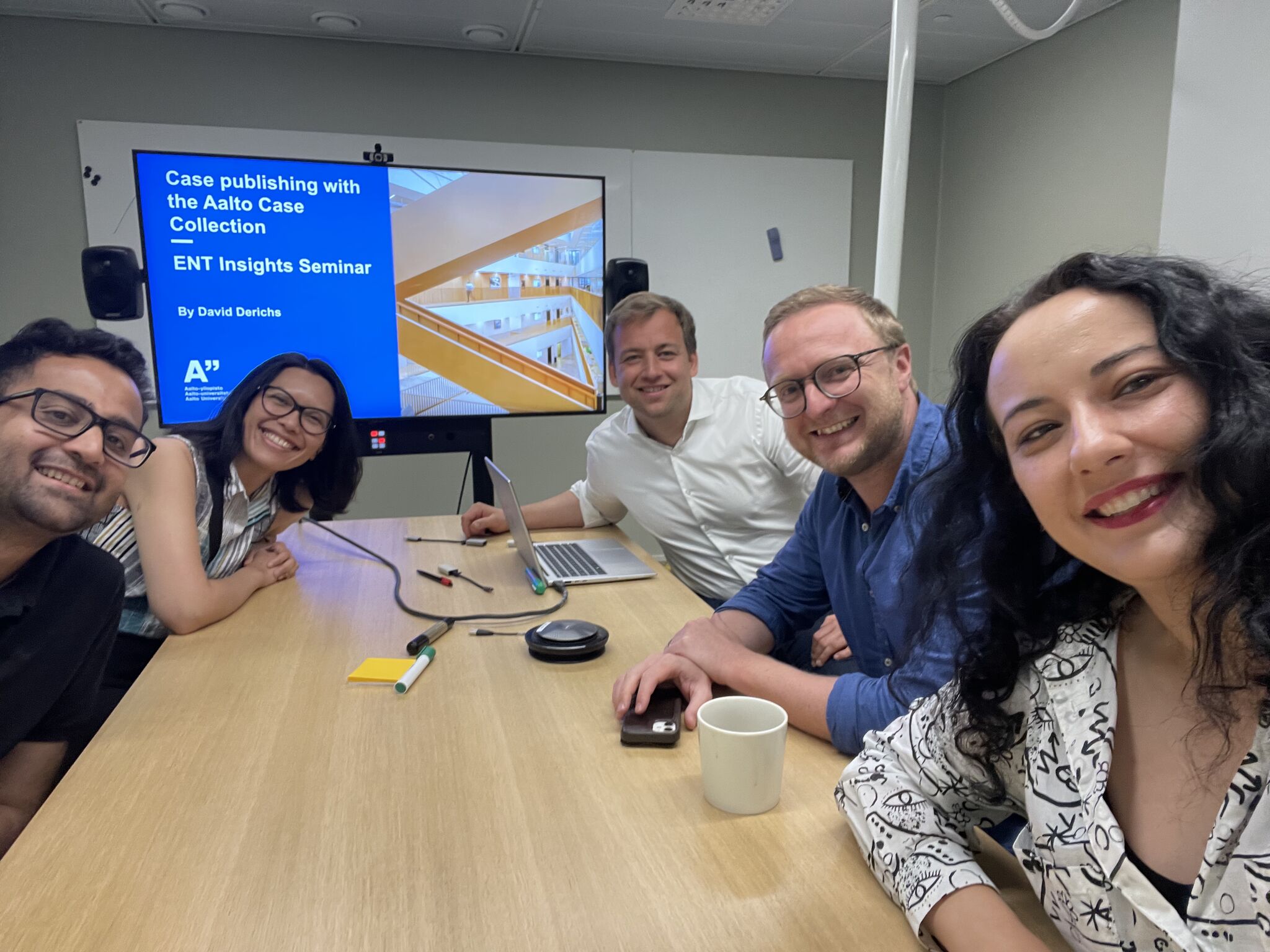 Five professionals seated around a conference table, smiling and looking at the camera. A presentation on a screen displays the title 'Case publishing with the Aalto Case Collection' at an ENT Insights Seminar.