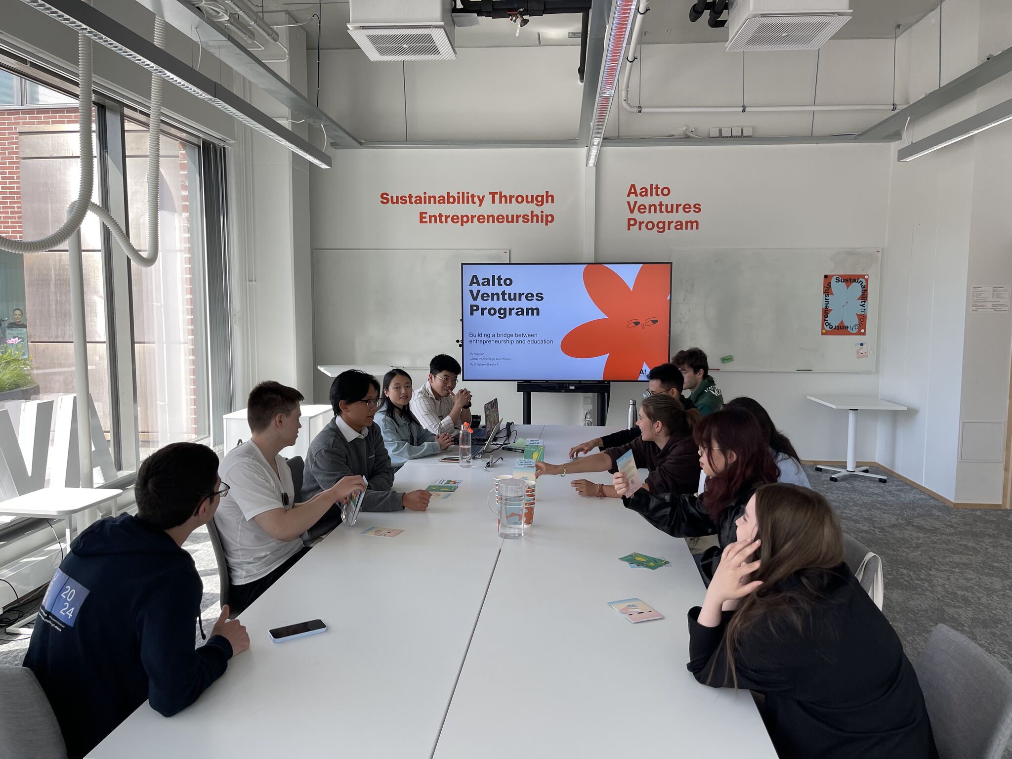 A group of people seated around a long white table in a modern meeting room, discussing topics related to entrepreneurship. A large screen displays the Aalto Ventures Program logo and a floral graphic. Bright sunlight illuminates the space.