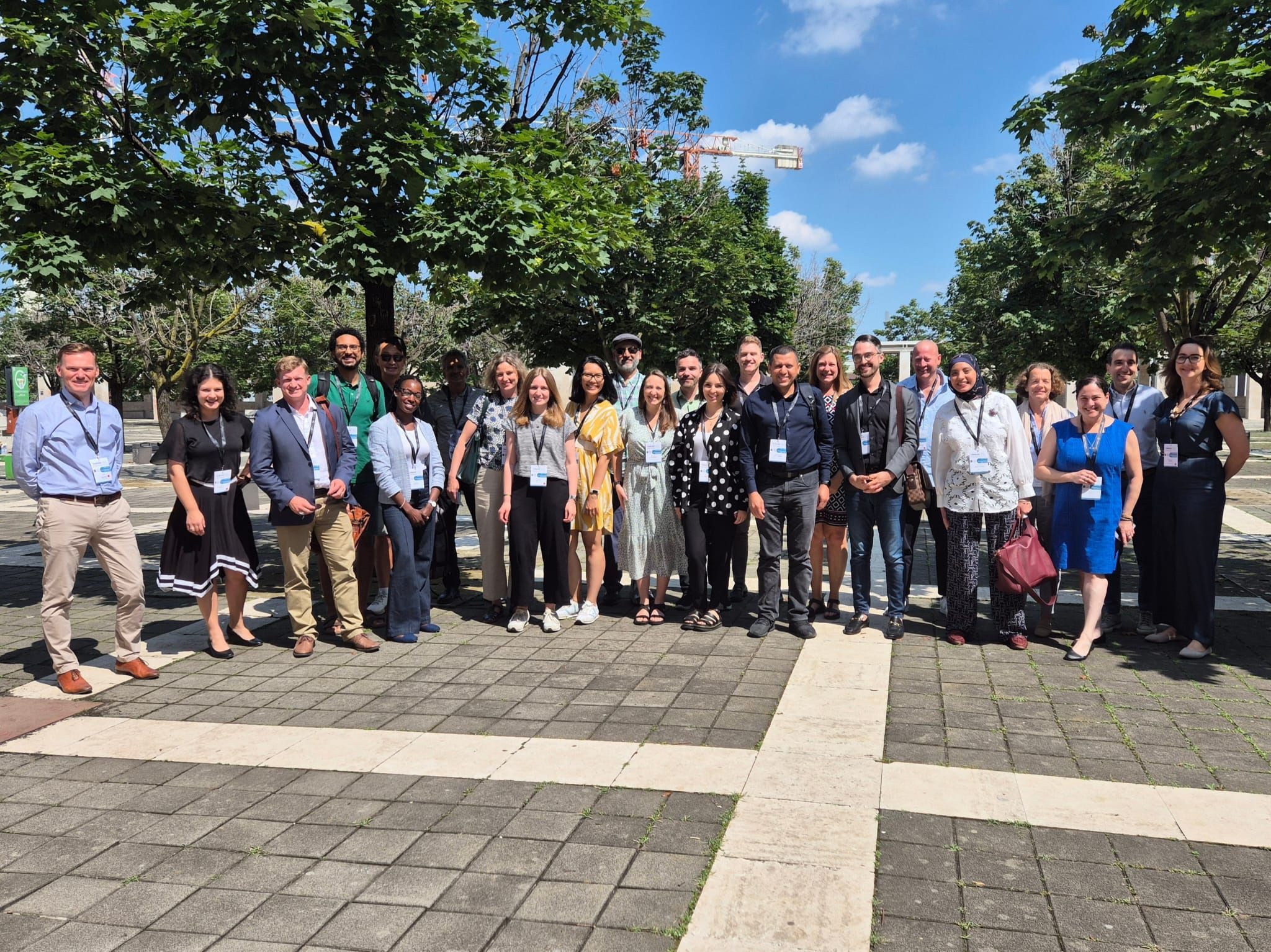 A group photo of approximately twenty people standing together in a sunny outdoor setting, surrounded by green trees. They are smiling and wearing name tags, with a mix of casual and formal clothing.