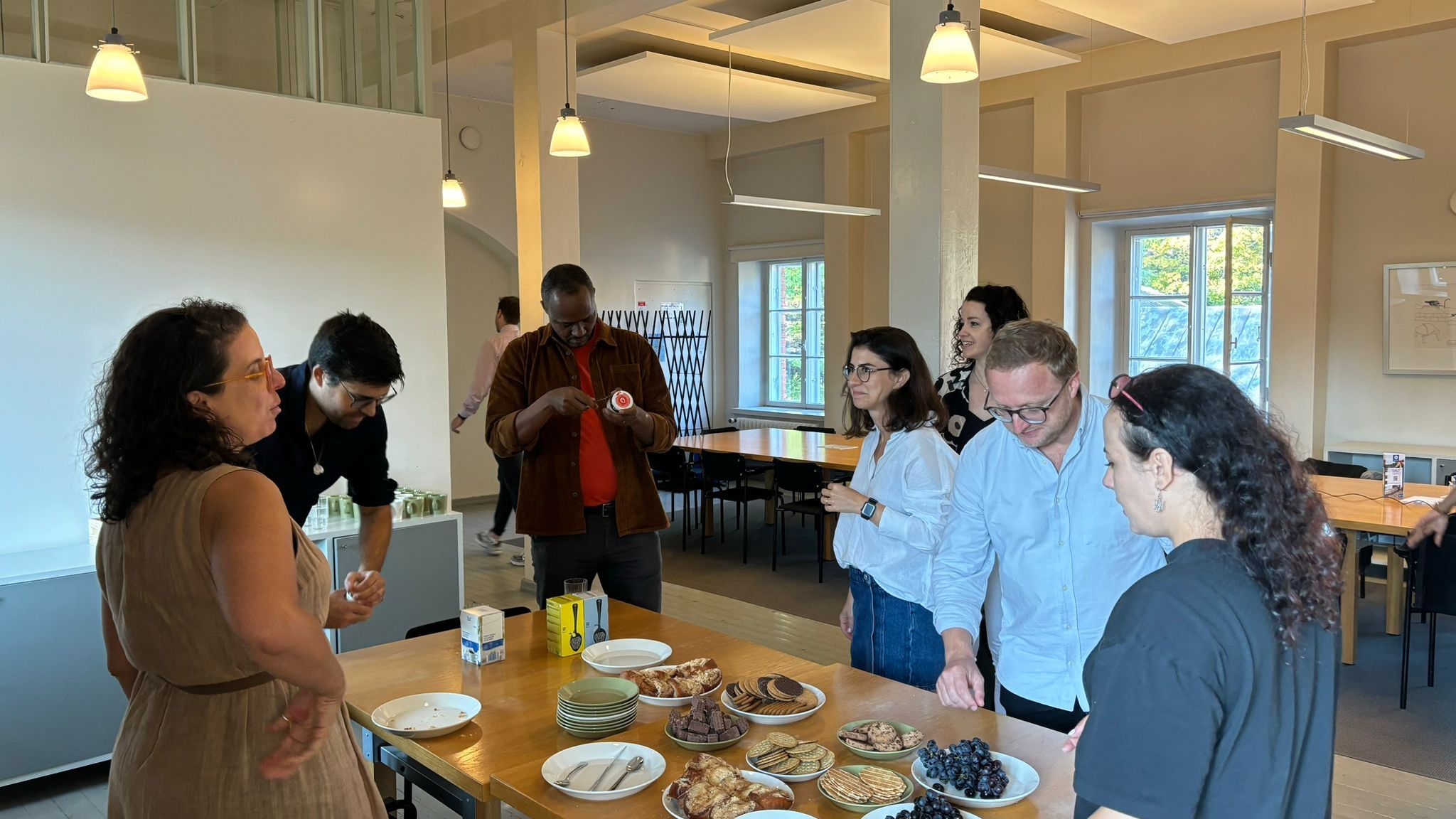 A group of six people engaged in conversation around a table filled with various foods, including cookies and fruits, in a bright, spacious room with large windows. They are casually dressed and appear to be enjoying a social gathering.