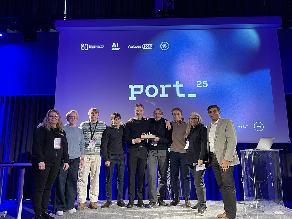 A group of eight people standing together on stage at a conference, smiling and holding an award in front of a colorful backdrop displaying the text 'port_ 25'. The stage is illuminated with blue and purple lights.