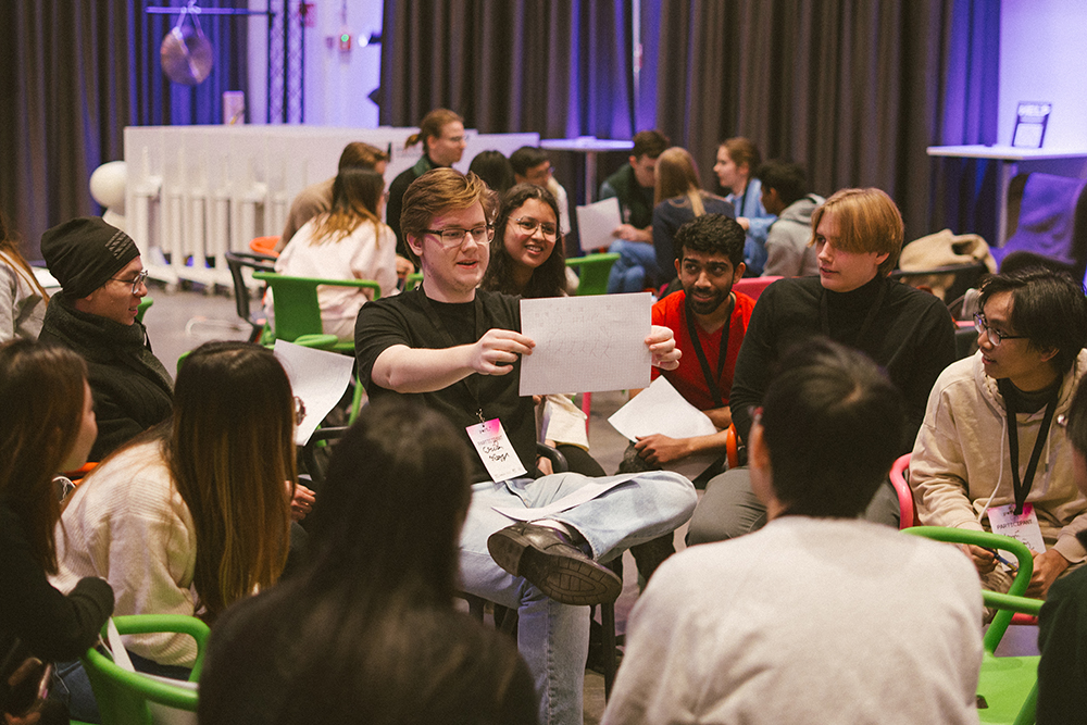 A group of diverse young adults seated in a circle, engaging in a discussion. One person in the center holds a piece of paper while others attentively listen, with a mix of expressions reflecting curiosity and interest.