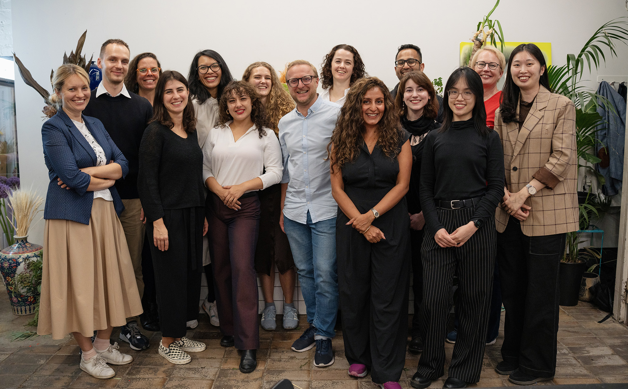 A diverse group of 15 people posing for a photo in a bright room with greenery. They are dressed in a mix of professional and casual attire, smiling and standing closely together, creating a friendly and inclusive atmosphere.