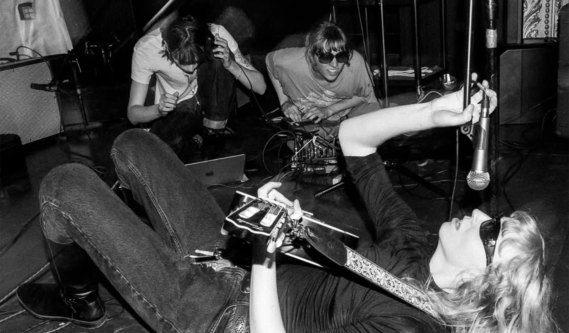 Black-and-white photo of three musicians on a stage. One person lies on the floor playing guitar while holding a microphone above their face. Two others crouch nearby adjusting audio equipment. All appear relaxed and immersed in the performance.